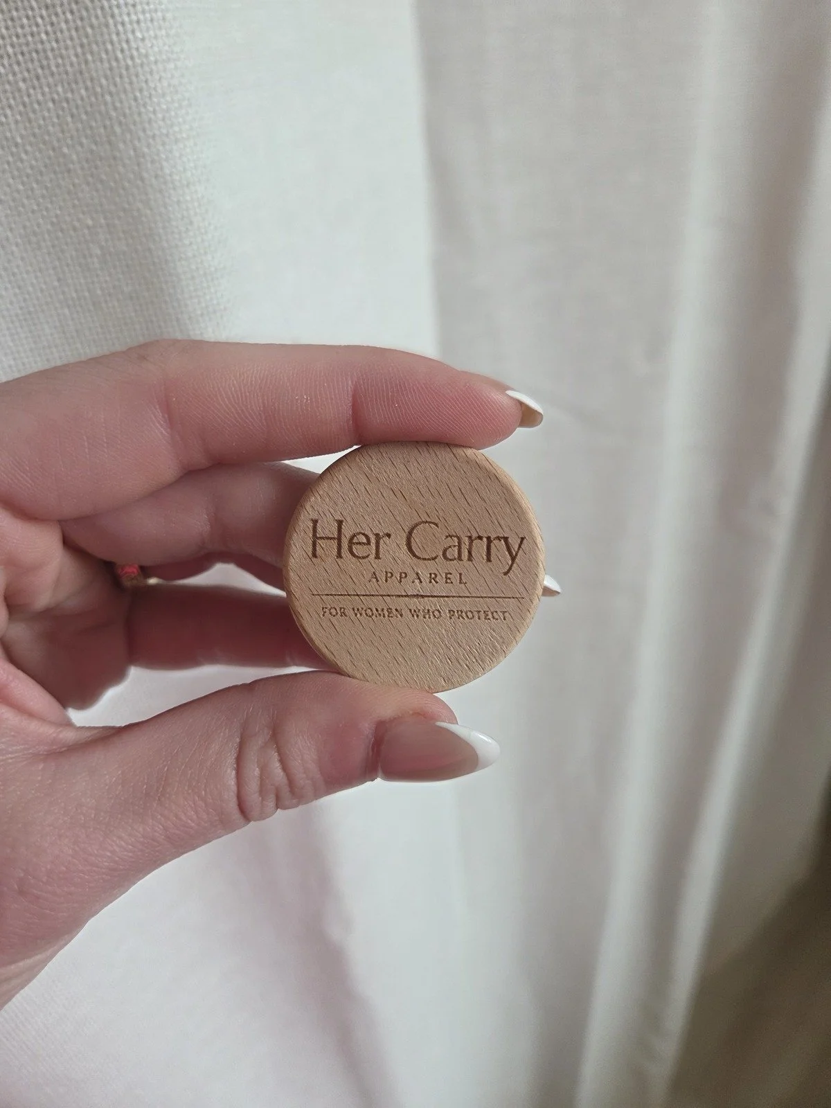 A hand holding a round wooden container labeled 'Her Carry Apparel' with the subtitle 'For Women Who Protect' against a plain background.