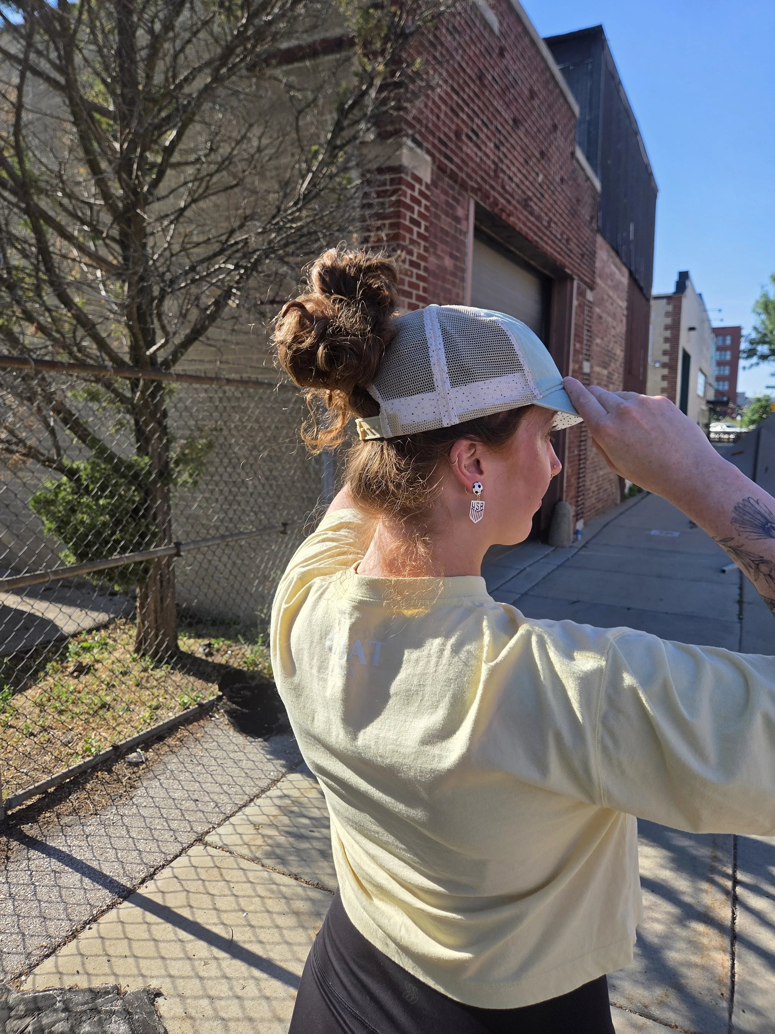 Woman wearing a ball cap supporting Carry It Forward mission. Her hair is in a curly bun through the ponytail opening in the hat.