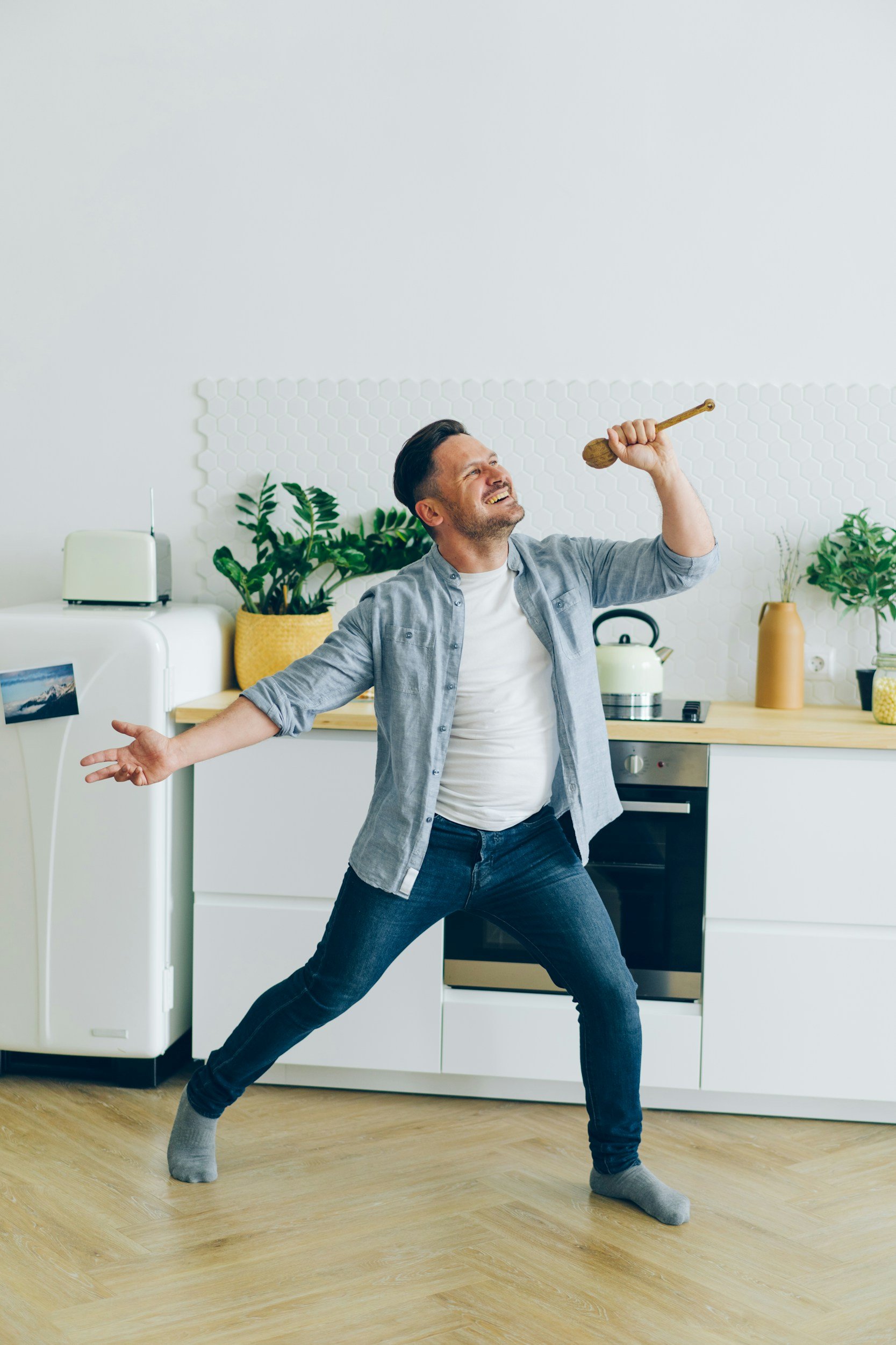 A man dancing in a kitchen with his arms and legs spread wide, holding a wooden spoon in one hand, wearing a gray shirt, jeans, and gray socks, with plants and kitchen appliances in the background.