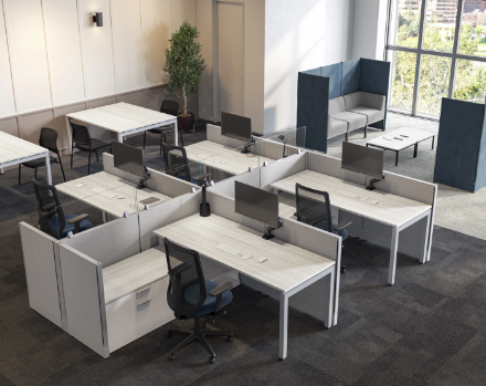 Empty modern office space with six white desks arranged in a U-shape, each equipped with black office chairs, desktop computers, and some with headsets. There is a separation wall between desks and a sitting area with a high-backed blue and gray lounge chair by a large window with city views, decorated with a tall potted plant.