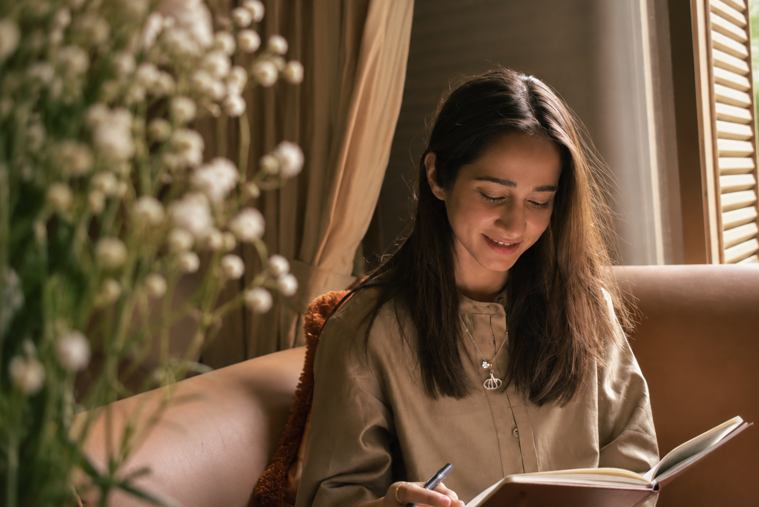A young woman with long dark hair sitting on a beige sofa near a window with wooden shutters, smiling and writing in a notebook, with a blurred bouquet of small white flowers in the foreground.