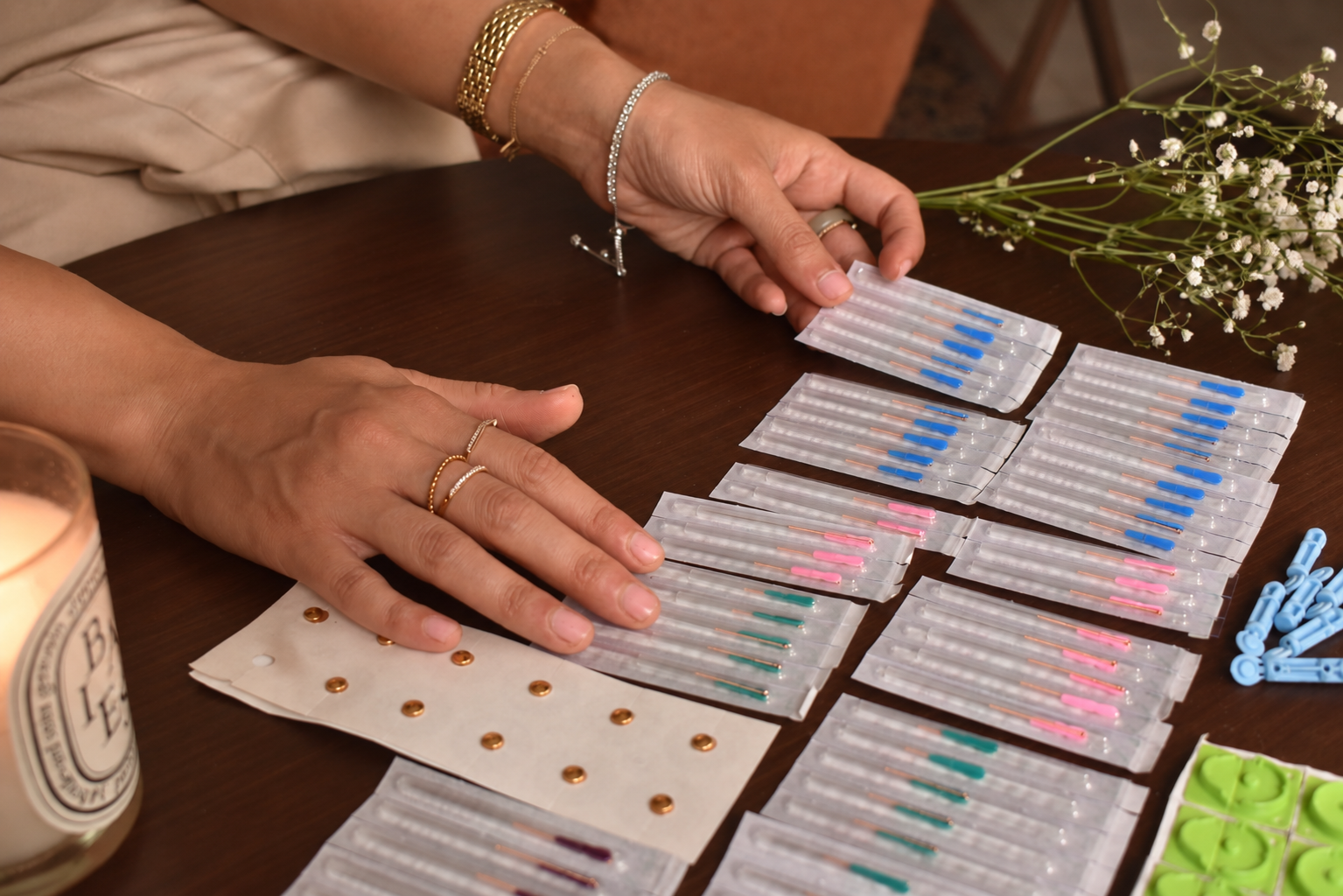 Hands organizing multiple packs of colorful test strips and pipettes on a wooden table, with a candle and flowers nearby.