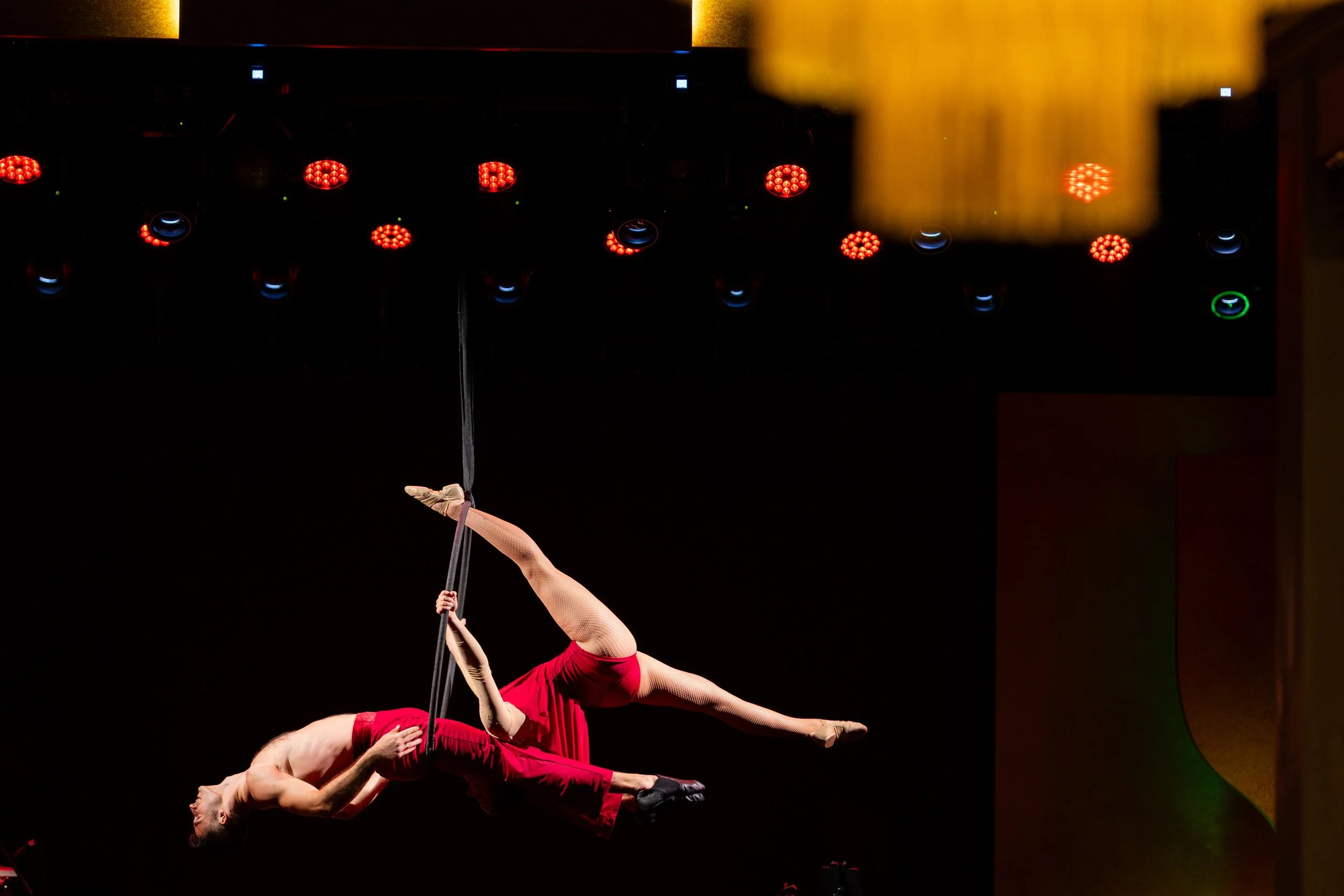 Aerial performers dressed in red costumes performing acrobatics on aerial silks against a dark background with stage lights.