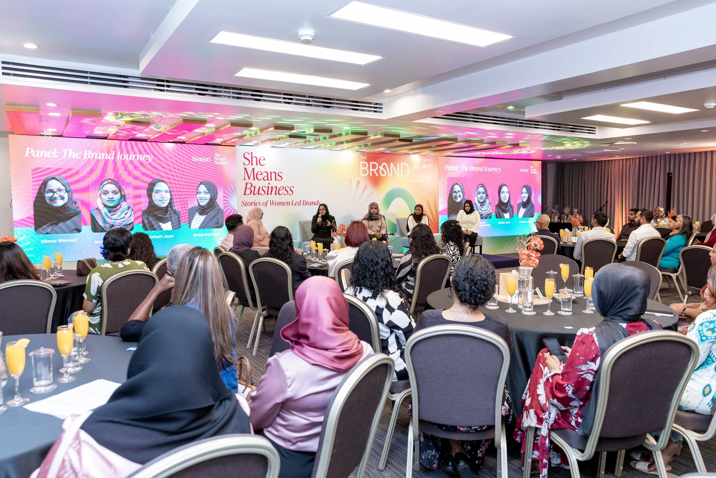 A large conference room with an audience listening to a panel discussion titled 'The Brand Journey.' The stage features a colorful backdrop with images of women and text about women-led brands, flanked by microphones, and rows of tables with attendees seated, many wearing headscarves and colorful clothing. There are drinks and notebooks on the tables.
