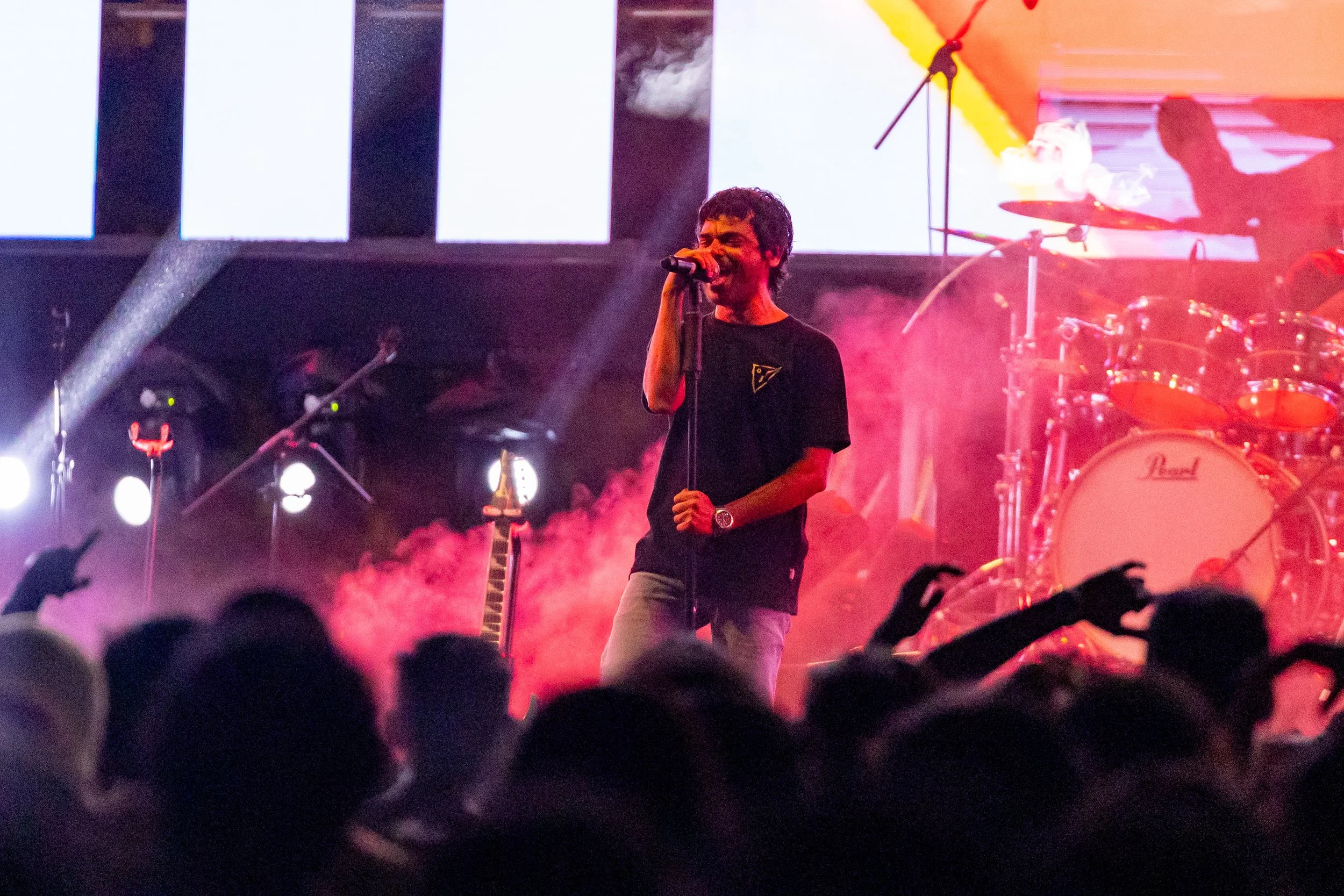 A male singer performs on stage with a microphone, illuminated by colorful stage lights and smoke effects. A drum set is visible in the background.