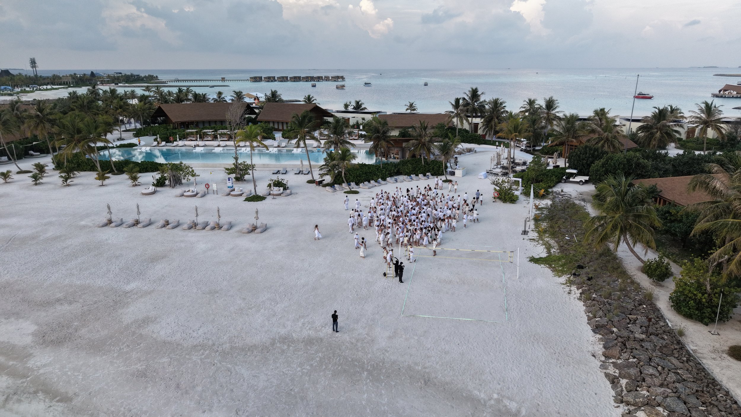Beach scene with a swimming pool, numerous lounge chairs and umbrellas, and a large group of people dressed in white gathered near a volleyball court. The background features palm trees, buildings, and a view of the ocean with boats.
