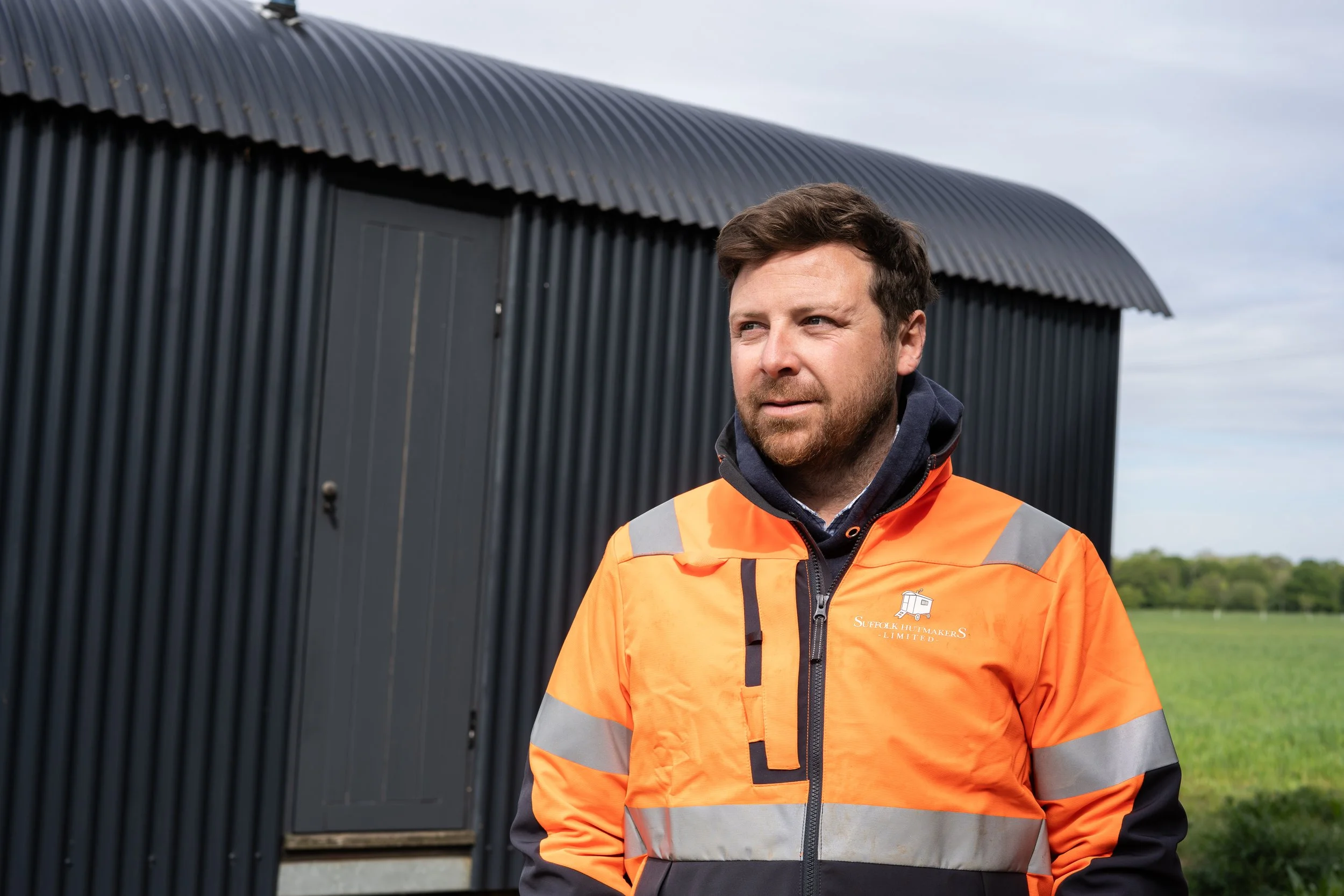 A man in an orange high-visibility jacket standing outdoors in front of a black corrugated metal building, with green field and trees in the background.