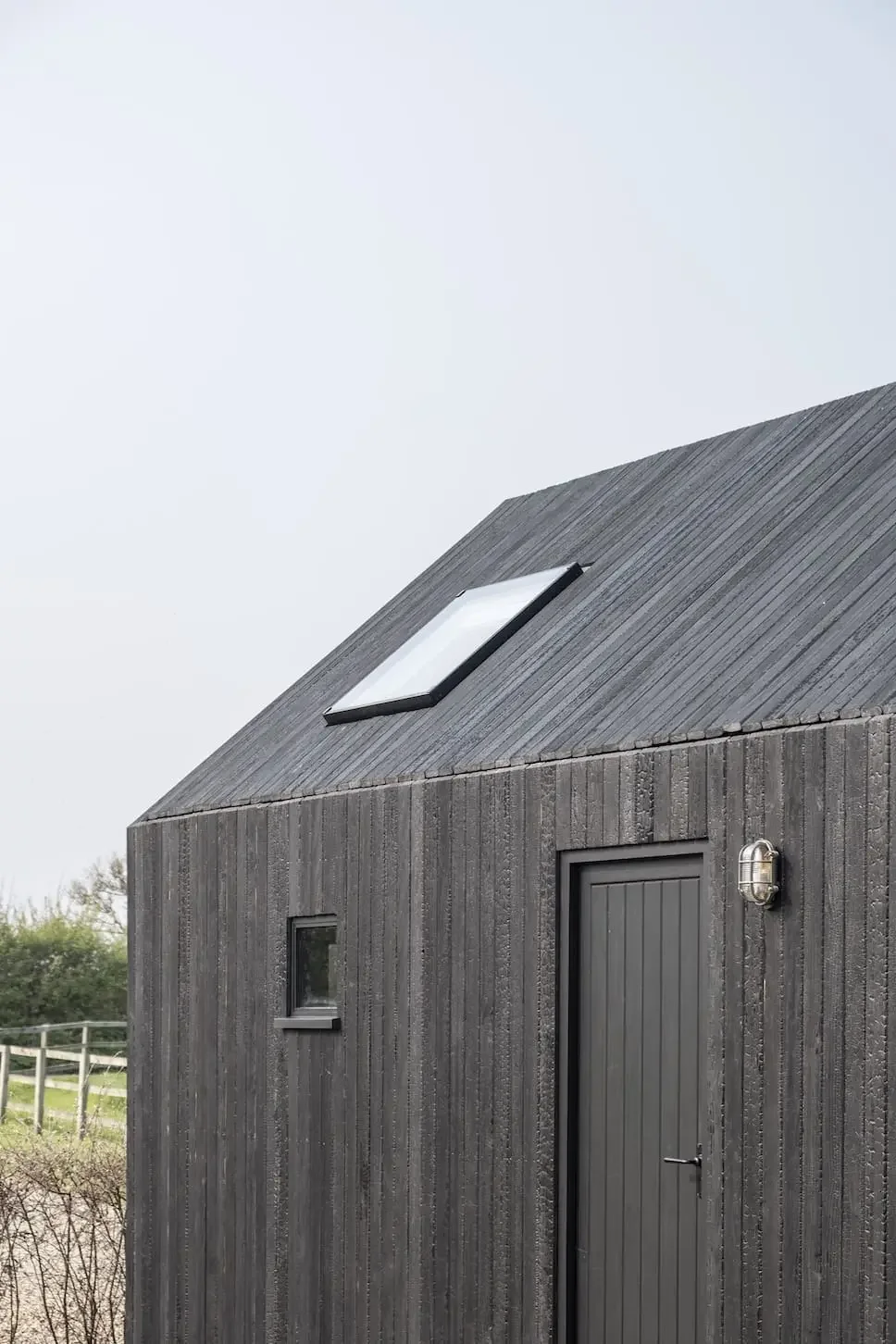 A small, dark gray barn or shed with vertical wooden siding, a single door, and a small window, featuring a skylight on the sloped roof and an exterior light fixture.