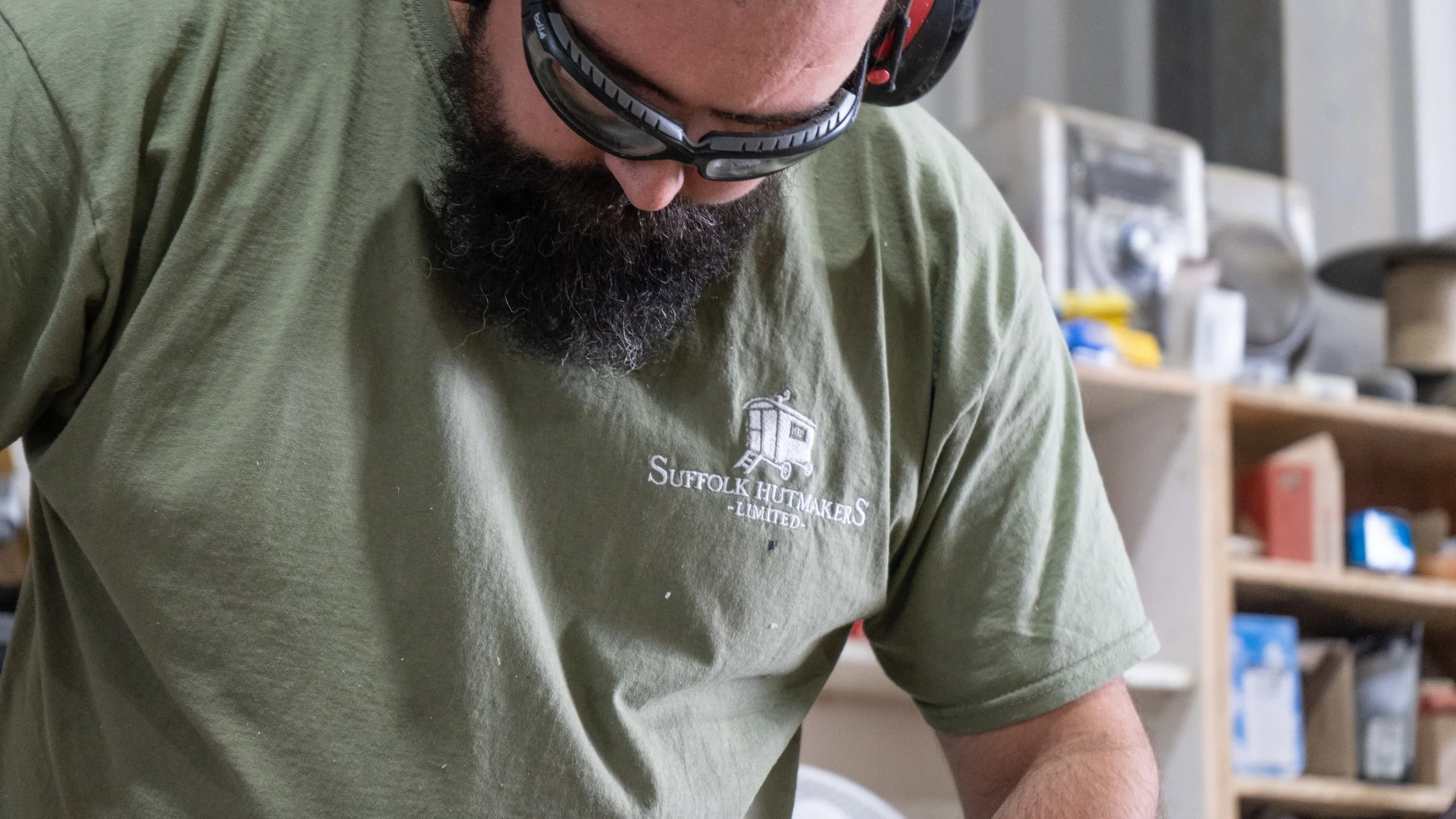 Close-up of a man with a beard wearing safety glasses and a green shirt with a logo that reads "Suffolk Hutmakers Limited," working in a workshop setting surrounded by tools and equipment.