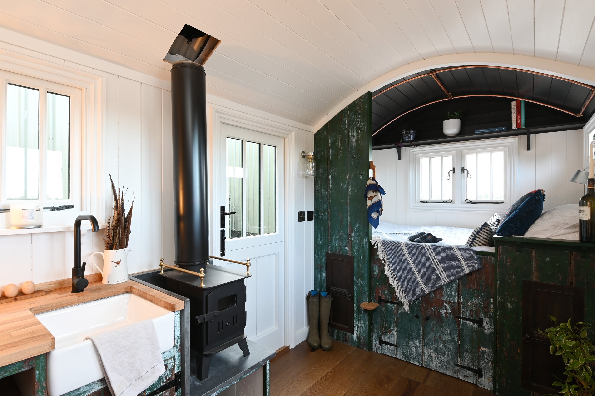 A cozy kitchen with white wooden panel walls, a black wood stove, and a view into a bedroom with windows, books, and plants.