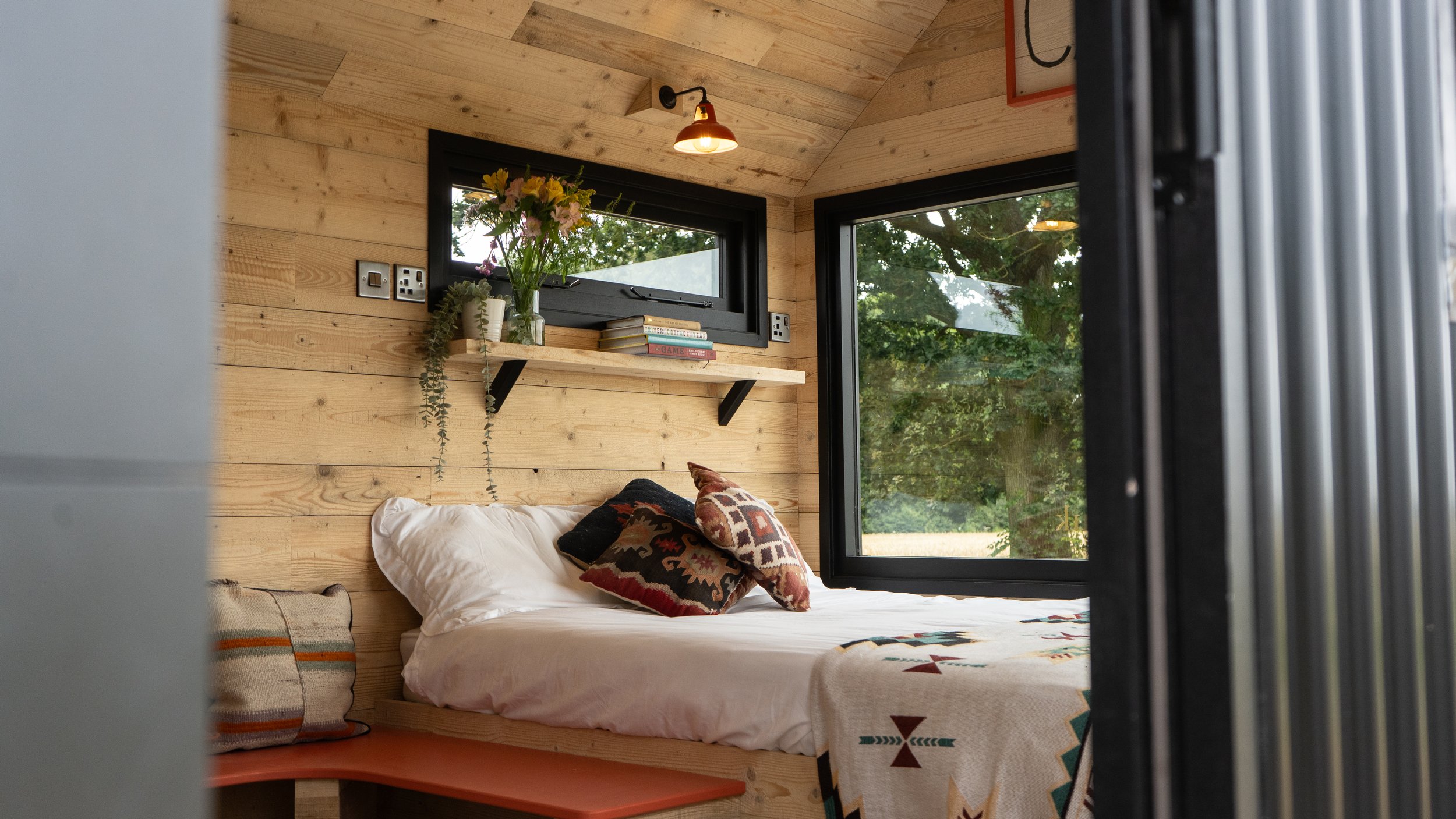 Interior of a cozy bedroom with wooden walls and ceiling, a bed with patterned pillows, a shelf with books and a vase of flowers, and large windows showing trees outside.