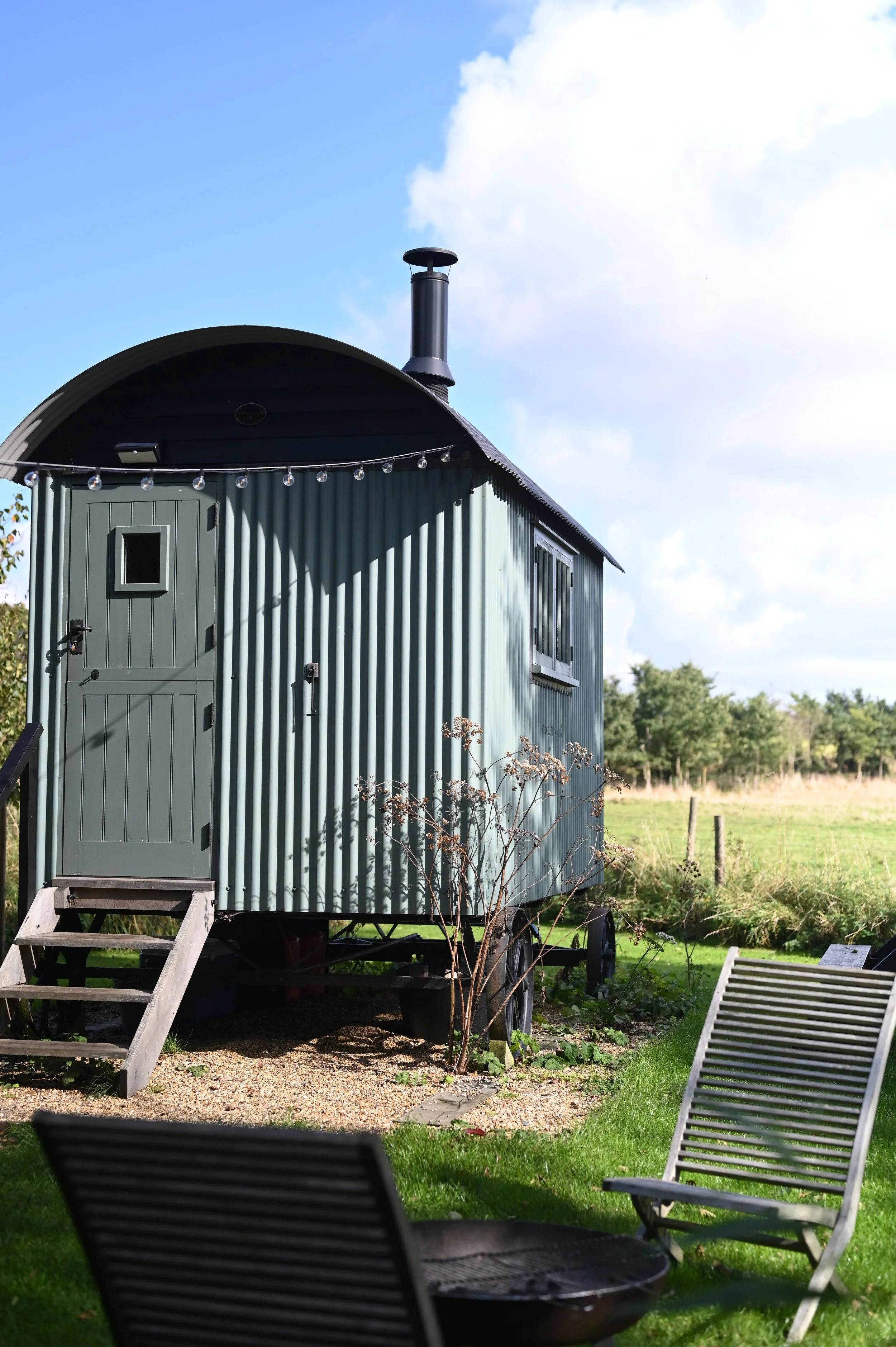 A small green metal building with a black chimney on a bright day, set against a blue sky with scattered white clouds, surrounded by greenery and trees.