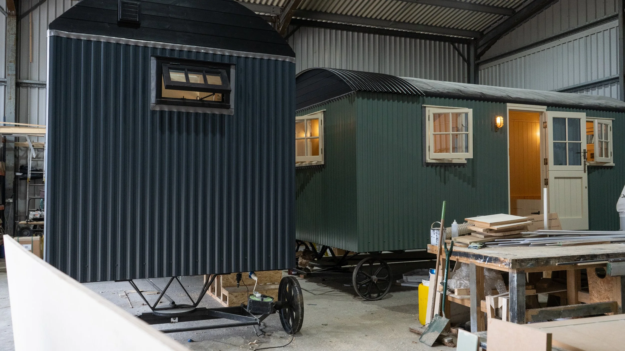 Two small, colorful houses with windows and doors inside a warehouse or workshop with metal walls and ceiling, surrounded by tools and materials.