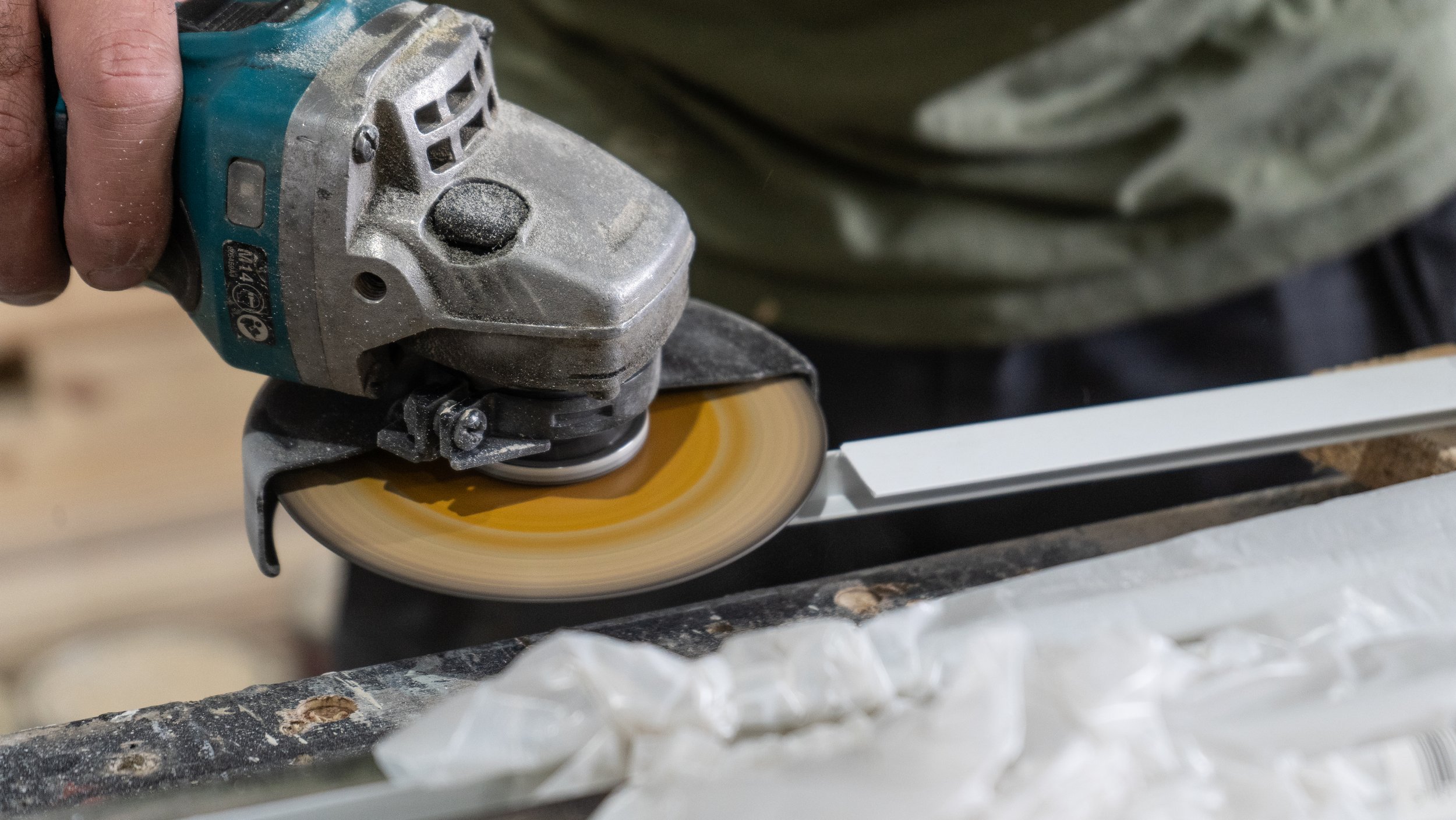 A person is using a handheld electric grinder to cut or polish a piece of wood or metal. The grinder is emitting dust or particles, and the person's hand and part of their clothing are visible.
