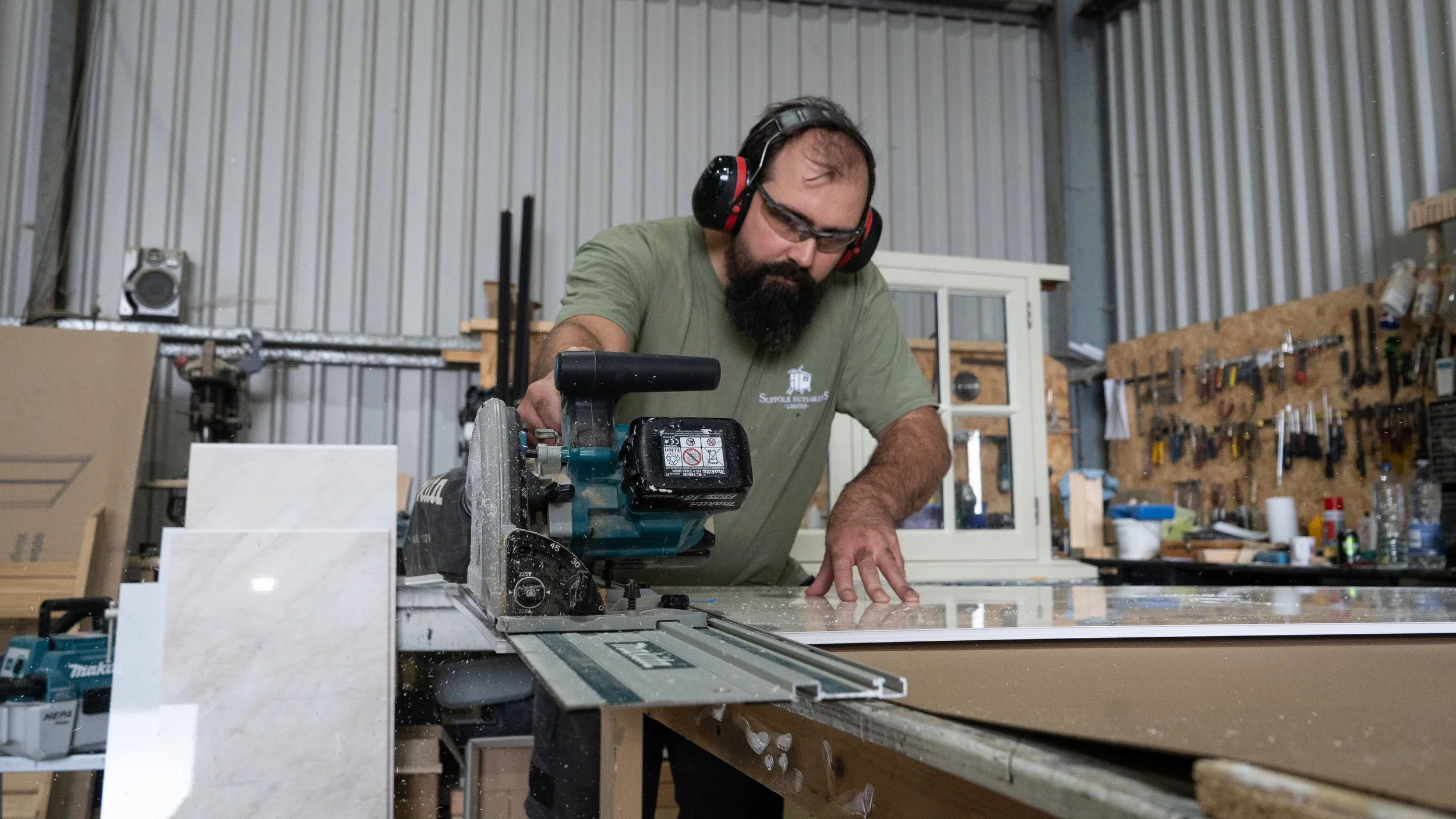 A man with a beard and safety glasses using a power saw in a woodworking shop, wearing ear protection and working on a piece of material on a workbench.