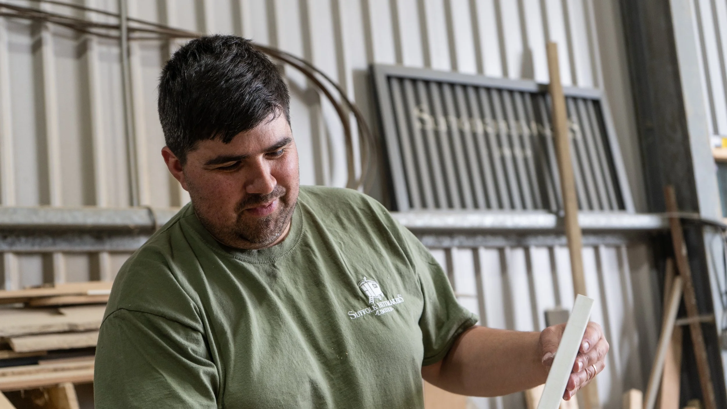 A man with short dark hair and a beard, wearing a green t-shirt, is looking down at a piece of wood or paper. He is indoors in a workshop or lumberyard, with wooden shelves and various pieces of wood in the background.