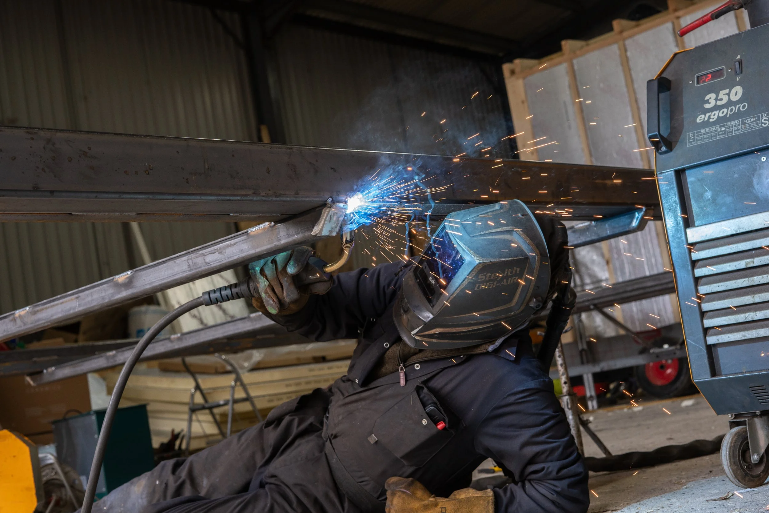 Worker welding metal beams in a workshop with sparks flying.