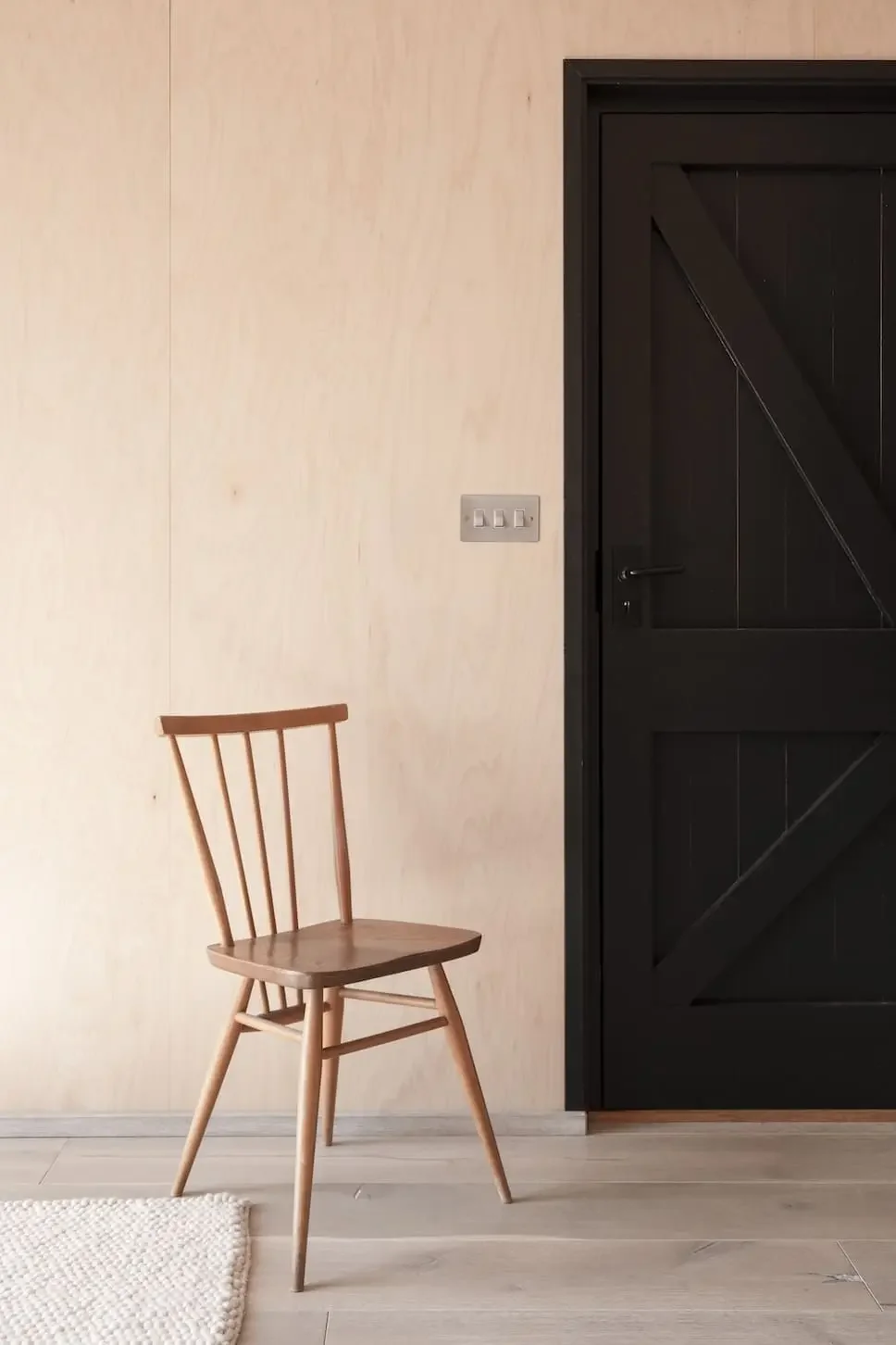 Empty wooden chair next to a black door in a room with beige walls and a light-colored floor.