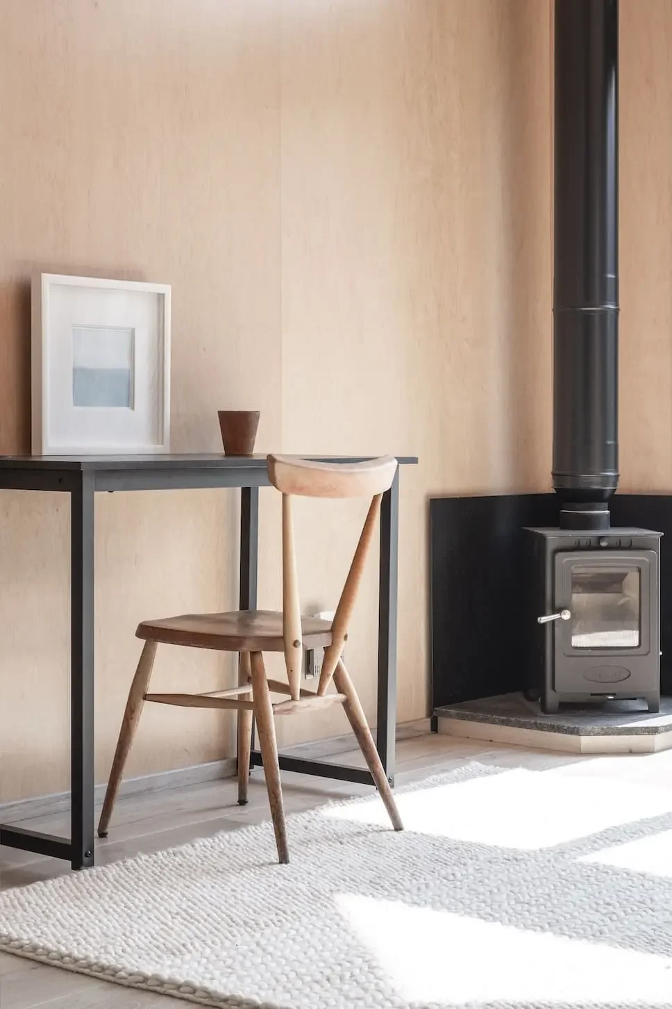 Minimalist interior with a wooden chair, a black desk with a framed picture and a small planter, and a black wood stove with a chimney against a beige wall, on a textured white rug.