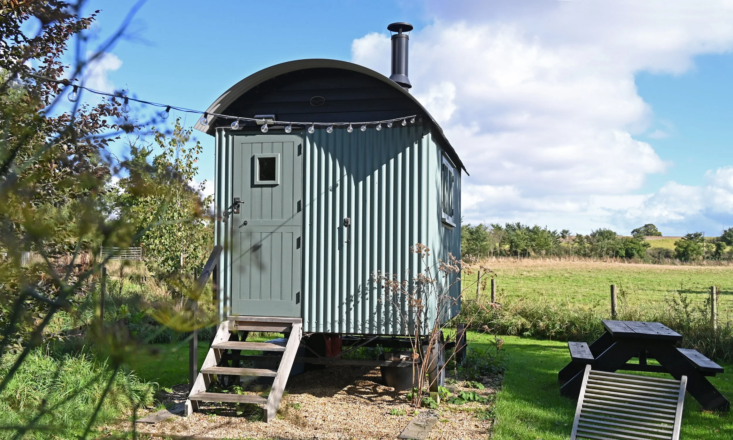 A tiny, blue, corrugated metal house with a rounded roof, a small door with a window, metal steps leading up to the door, and a skylight on the roof, set in a grassy outdoor area with trees and a cloudy sky.