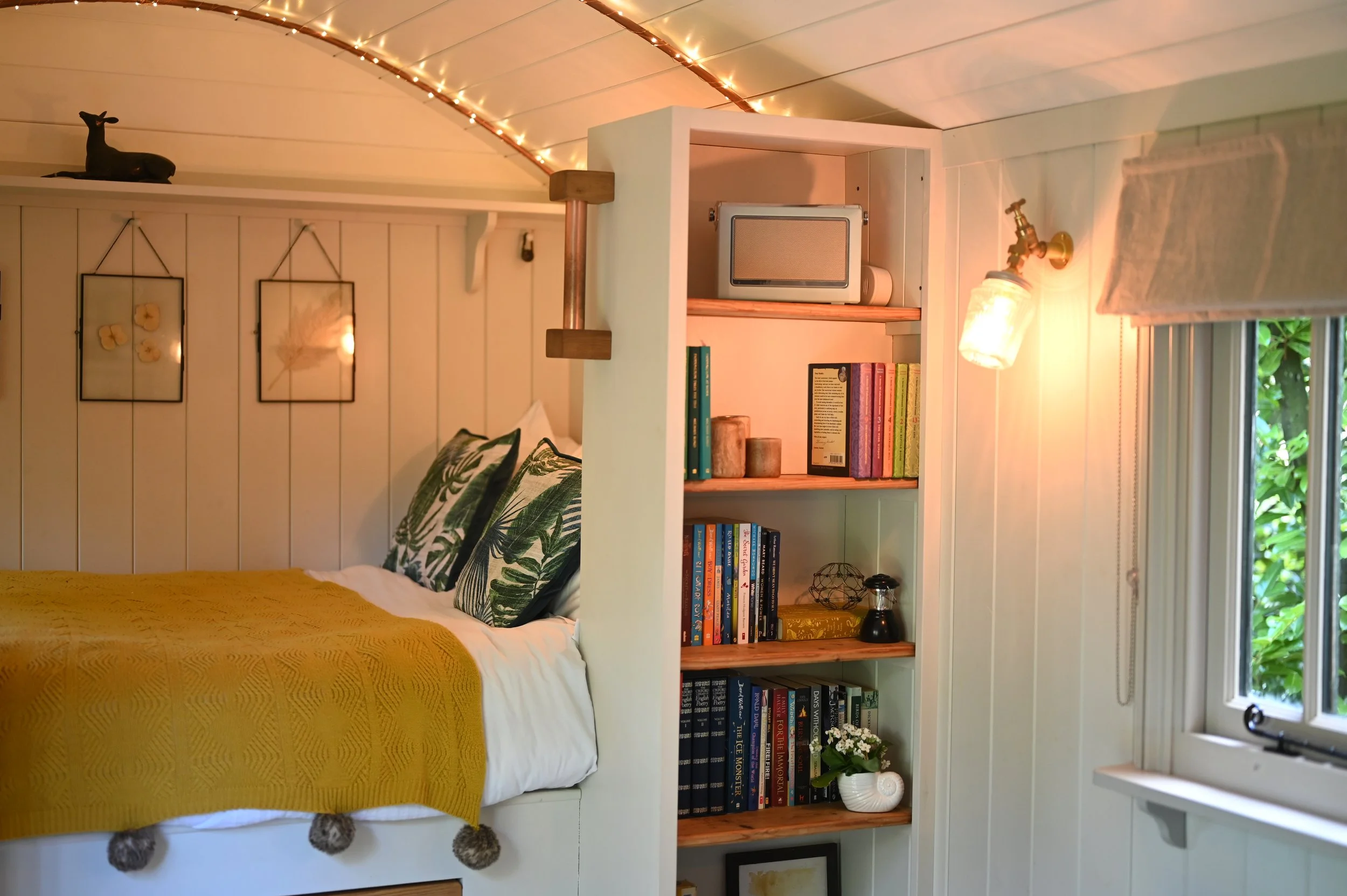 A cozy bedroom corner with a white bed featuring green leaf-patterned pillows and a yellow quilt, a white bookshelf filled with books, and warm lighting from string lights on the ceiling and a wall-mounted lamp near a window with a beige Roman shade 