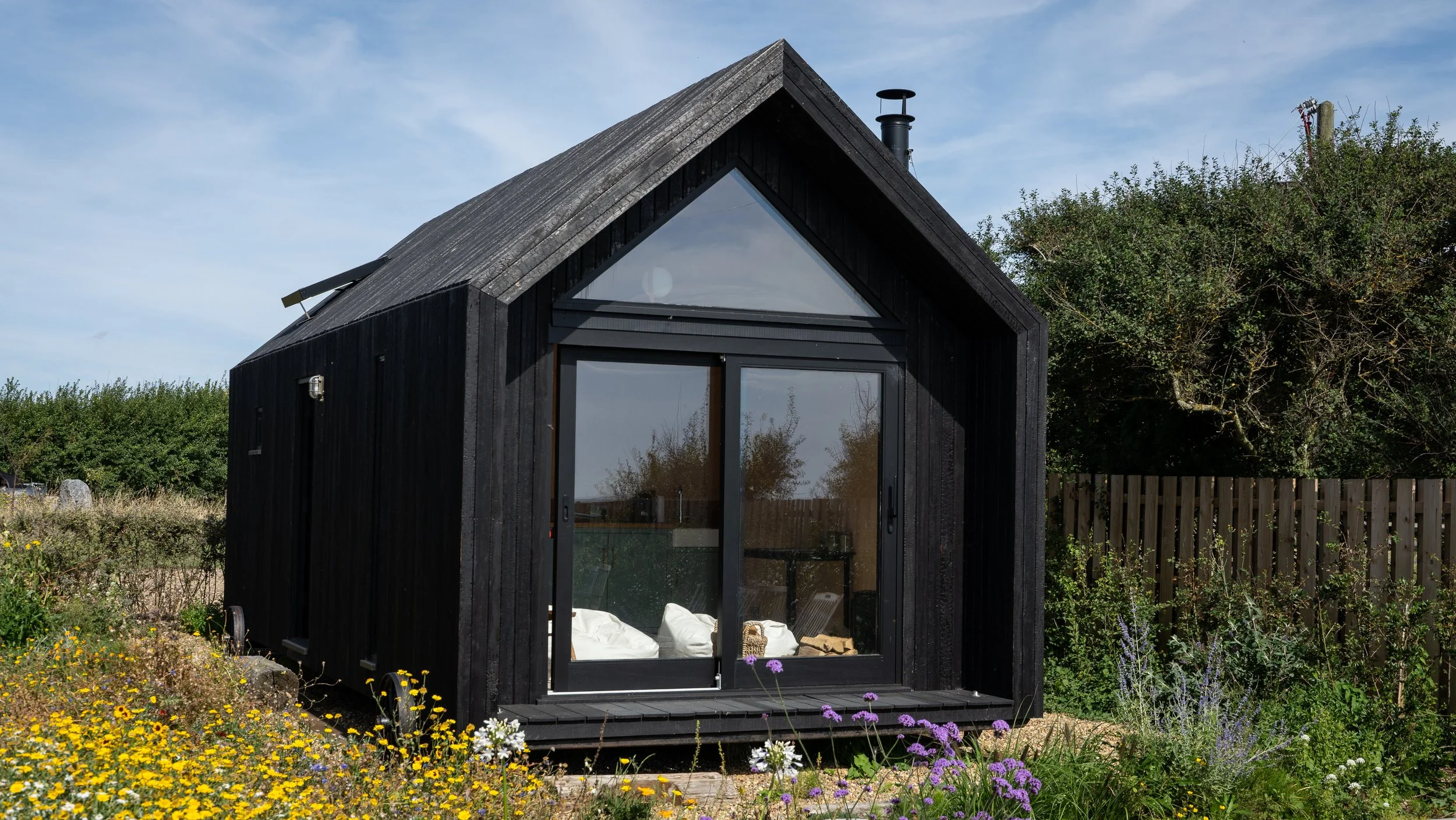 A small black house with large glass doors and a triangular window above, situated in a garden with colorful flowers and surrounded by a wooden fence, under a partly cloudy sky.