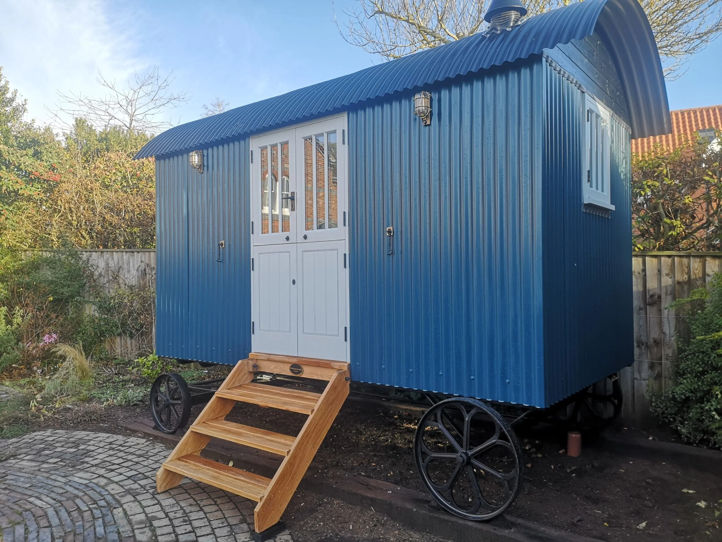 A small blue mobile tiny house on wheels with a white door and window, set outdoors with trees and a fence in the background. A wooden set of stairs leads up to the door.