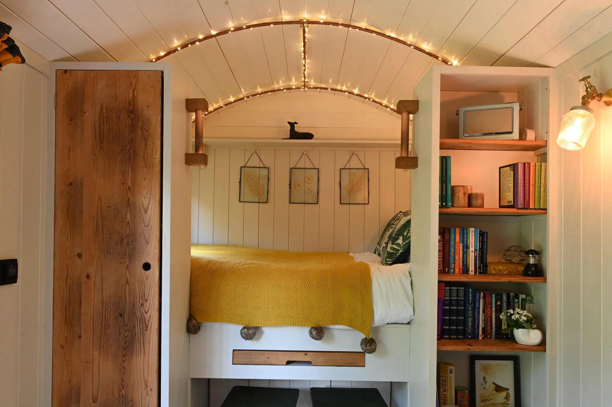 A cozy bedroom with a wooden bed frame, yellow bedding, and decorative pillows. The back wall is decorated with framed botanical prints. A bookshelf filled with colorful books and small decorative objects is on the right. String lights are draped acr