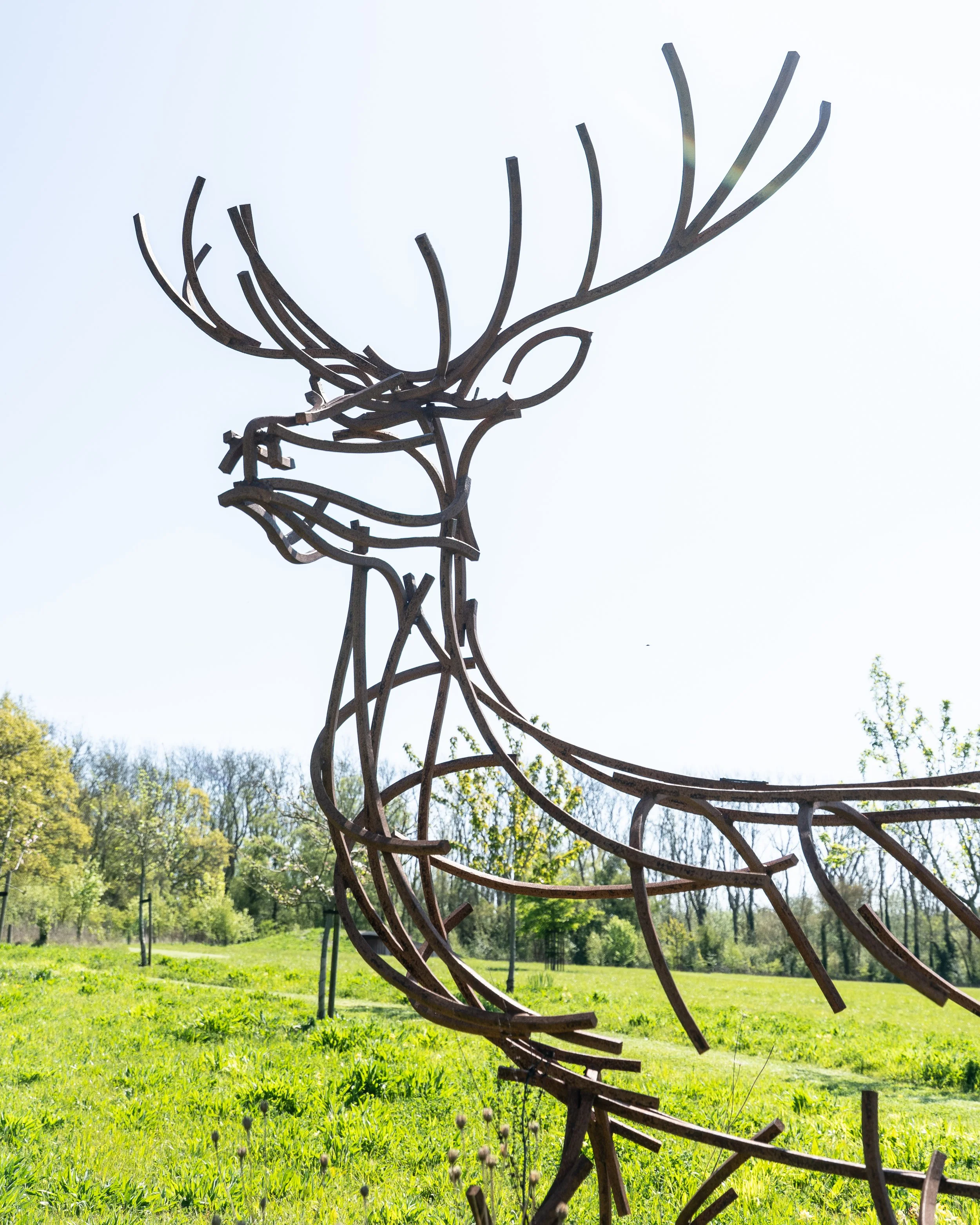 A deer-shaped metal sculpture in an outdoor park setting with green trees and grass in the background.