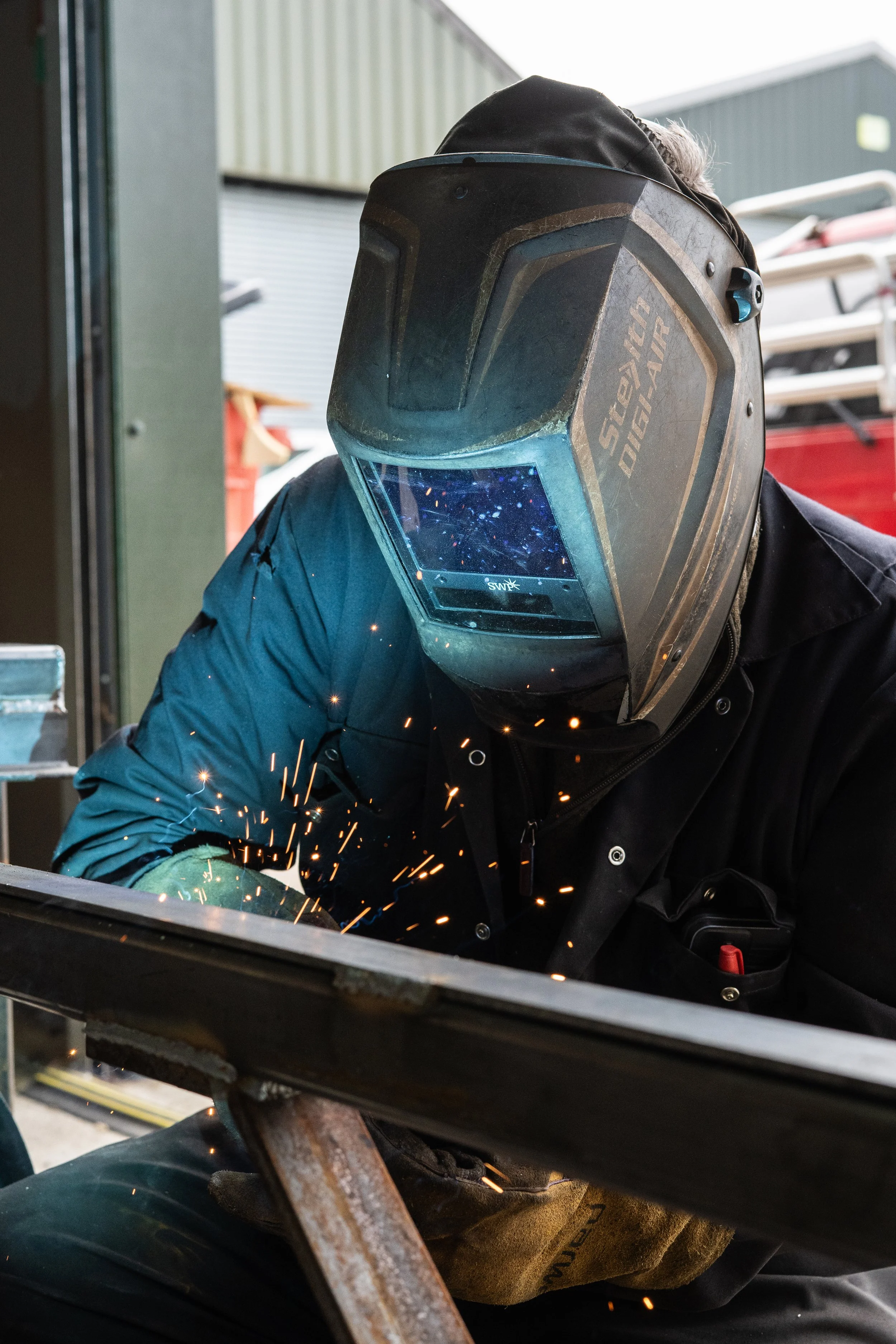 A person wearing a welding helmet and dark clothing welding metal, with sparks flying in a workshop or industrial setting.