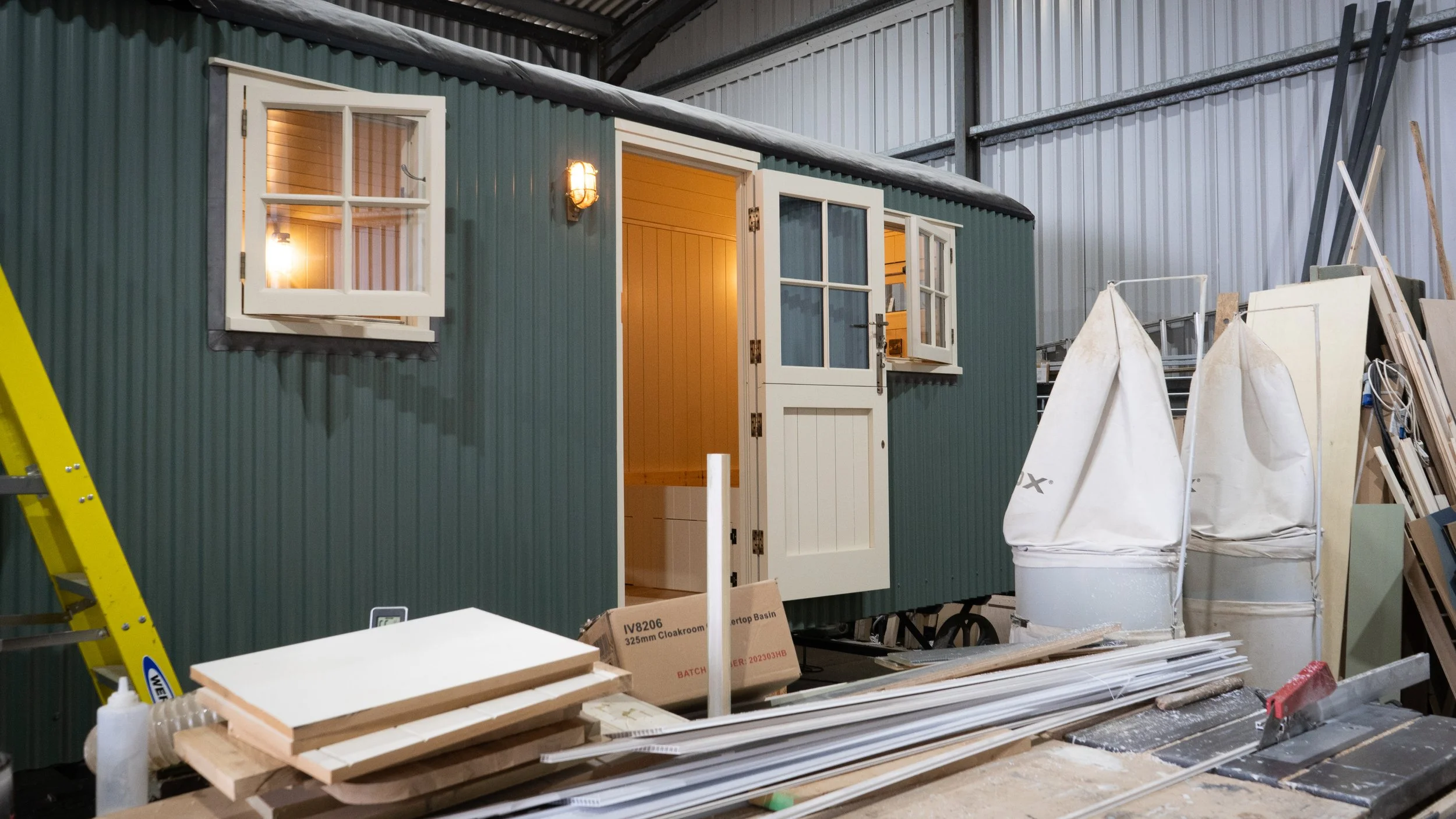 A miniature house with green exterior walls and white window and door frames is situated inside a warehouse. The house has a warm-lit interior visible through the open door and windows. Surrounding it are construction materials, boxes, and a yellow l