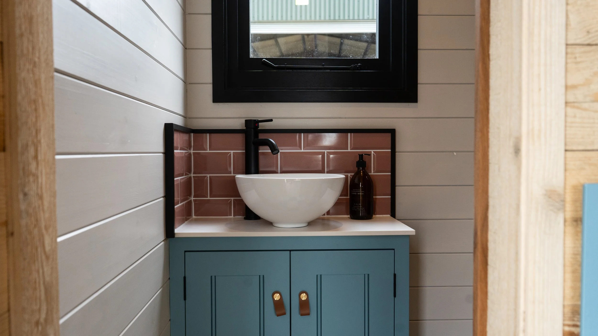 A small bathroom vanity with a white round vessel sink, black faucet, and a brown soap dispenser against a pink-tiled backsplash. There's a small window above the sink, framed in black, with a rain-covered outside view. The vanity is painted blue, and the walls are covered with white horizontal wooden planks.
