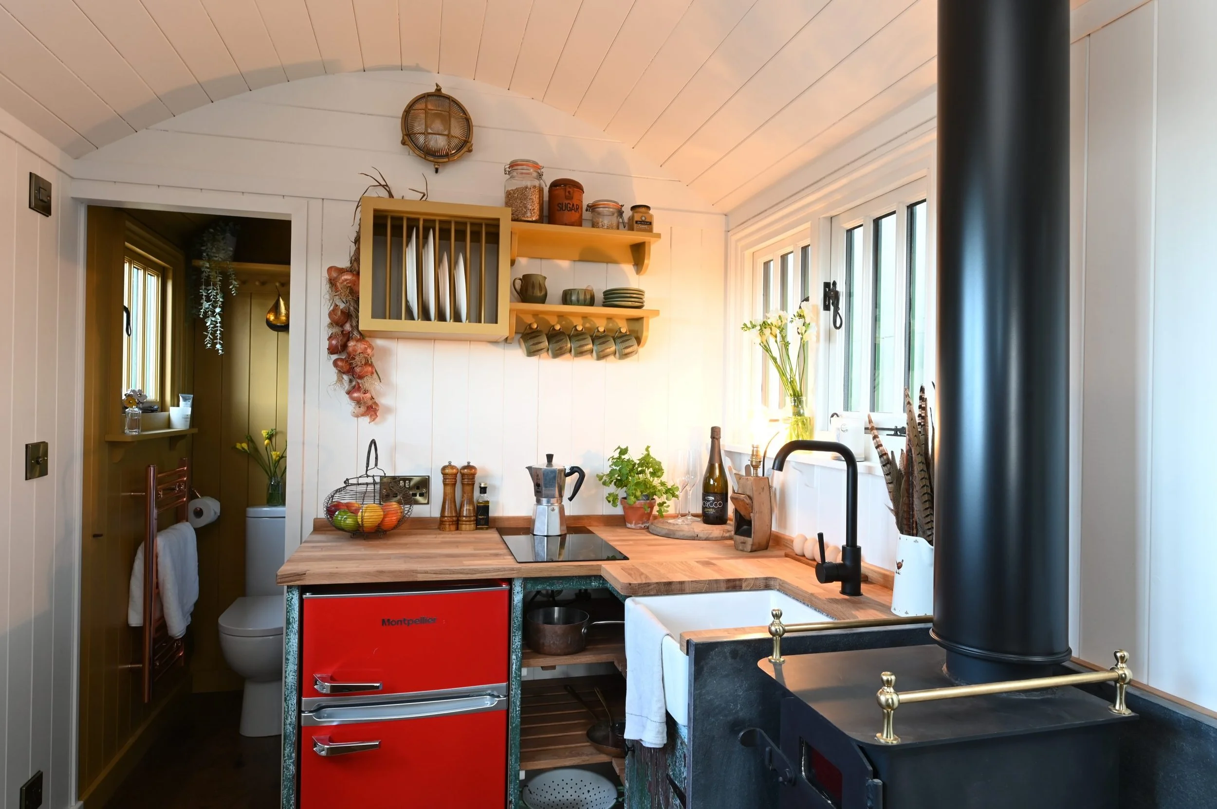 A cozy kitchen with white wood-paneled walls and ceiling. A black wood stove with brass accents is in the foreground. Wooden shelves hold plates and cups, and a jar collection is displayed above. A fruit basket, coffee grinder, and green plant decorate the countertop. Large windows let in natural light, and a small doorway reveals a toilet and laundry area.