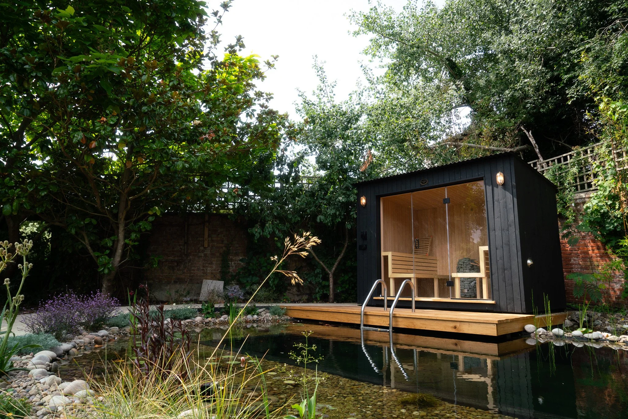 A backyard garden with lush green trees and plants, a small pond with rocks at the edge, and a modern black sauna with a glass door and wooden interior, situated on a wooden deck. Suffolk Sauna making.