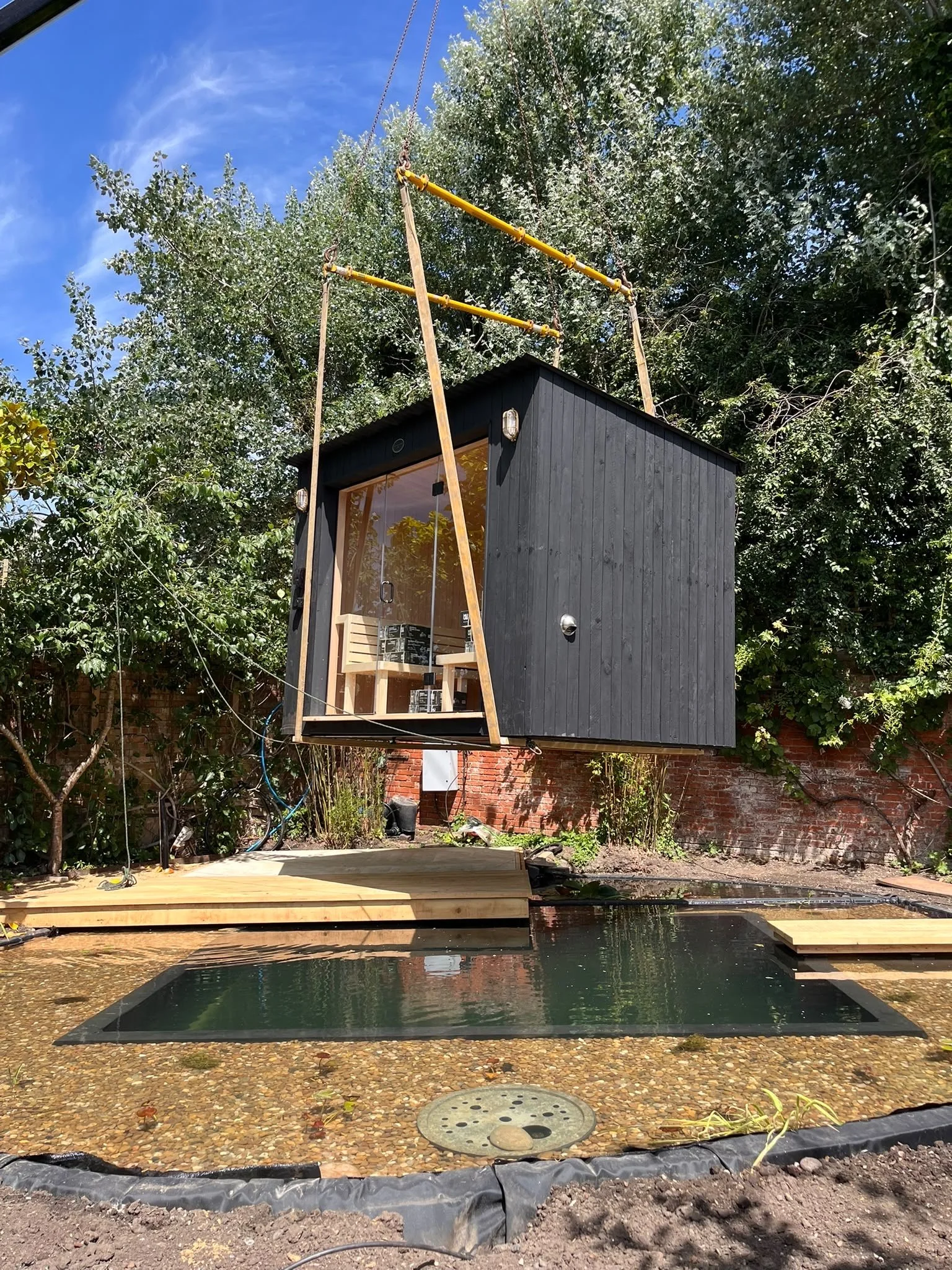 A small black wooden building with a glass door and an outdoor shower head, surrounded by trees and a brick wall. In the foreground, there is a pool with a pebble and tile bottom, and a wooden deck. The sky is clear with some clouds.