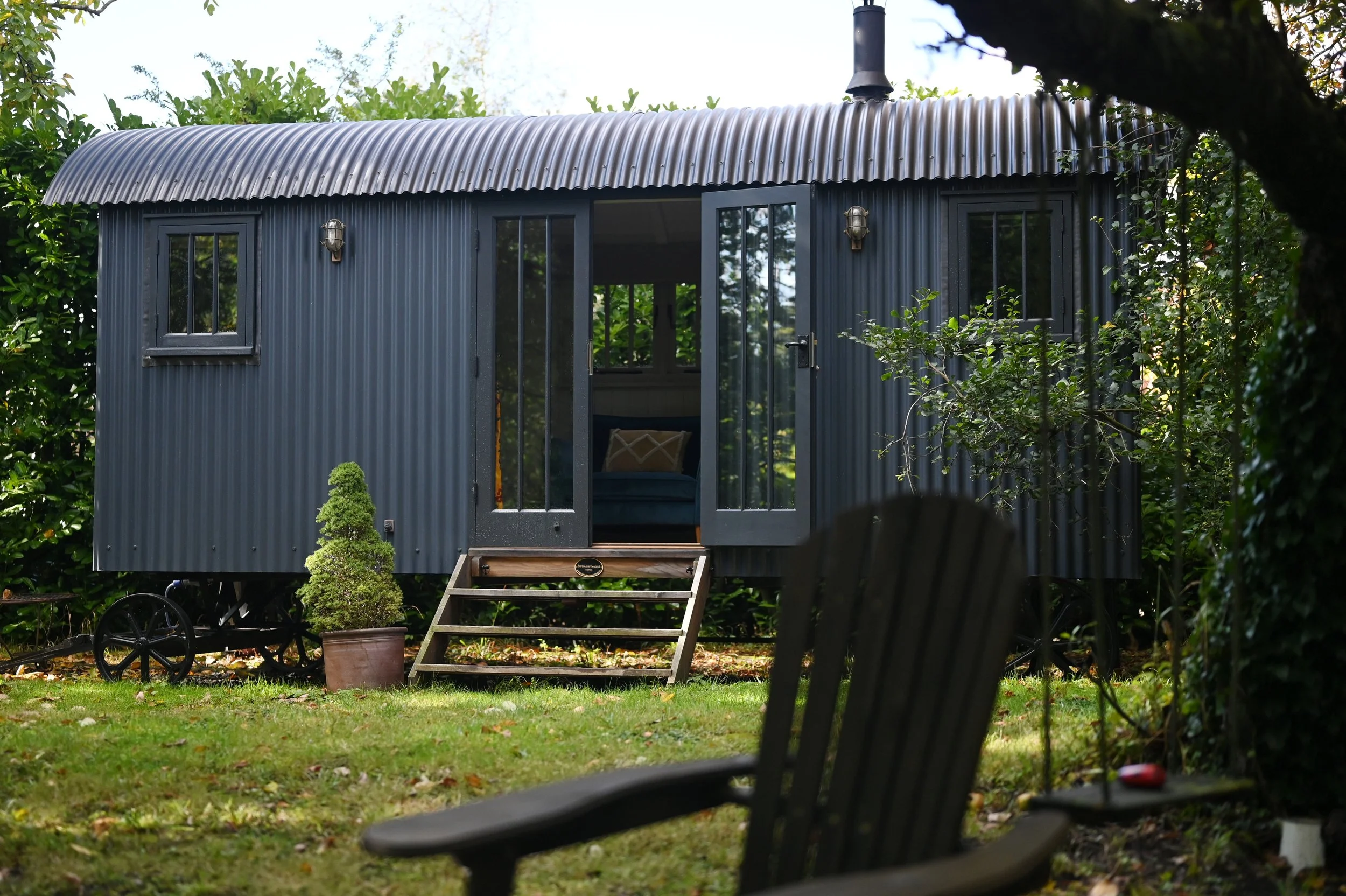A tiny gray metal cabin with two small windows and a glass door, surrounded by green trees and shrubs, with a small staircase leading to the entrance. In the foreground, part of a black metal garden bench and a potted plant are visible.
