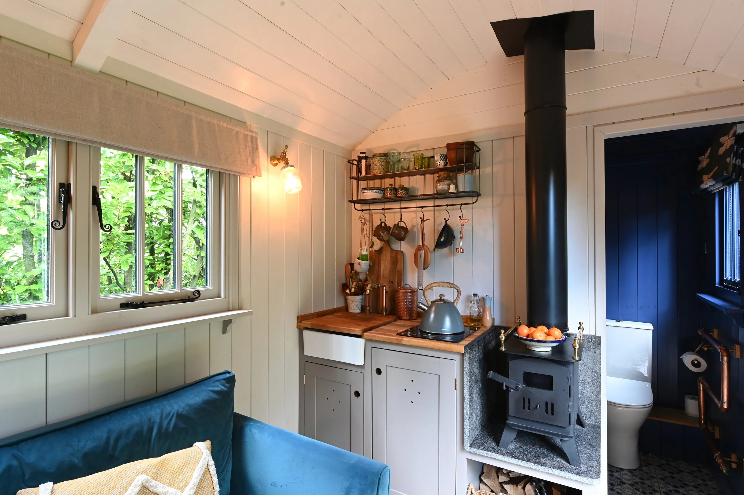 A cozy kitchen corner featuring a wooden countertop, wall-mounted shelves with jars and cups, hanging pots and utensils, a black stove pipe, a window with green foliage outside, and part of a blue couch with a yellow pillow.