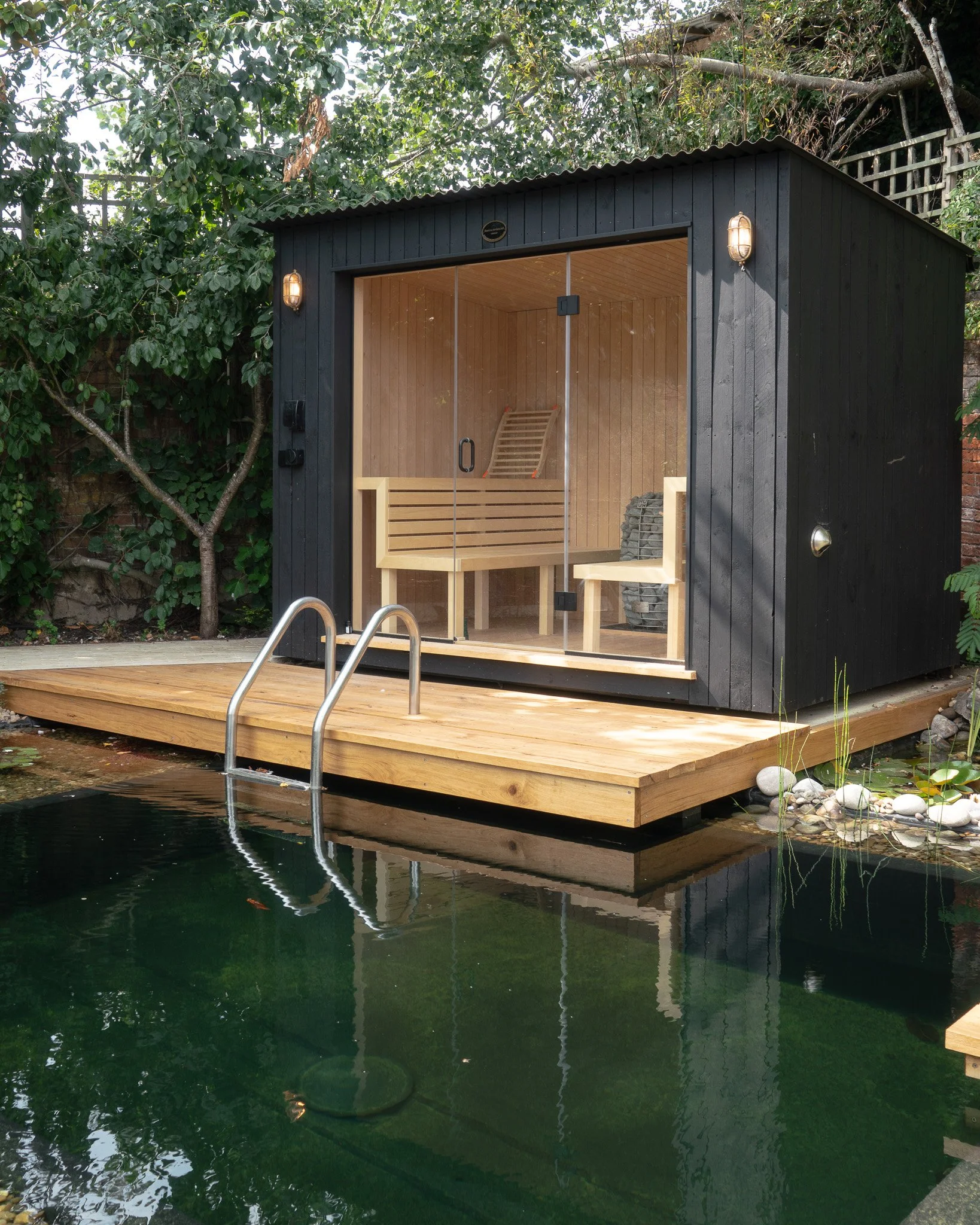 A small black wooden sauna with a glass front, situated on a wooden deck over a pond with green water, surrounded by greenery and rocks. Suffolk Sauna making.