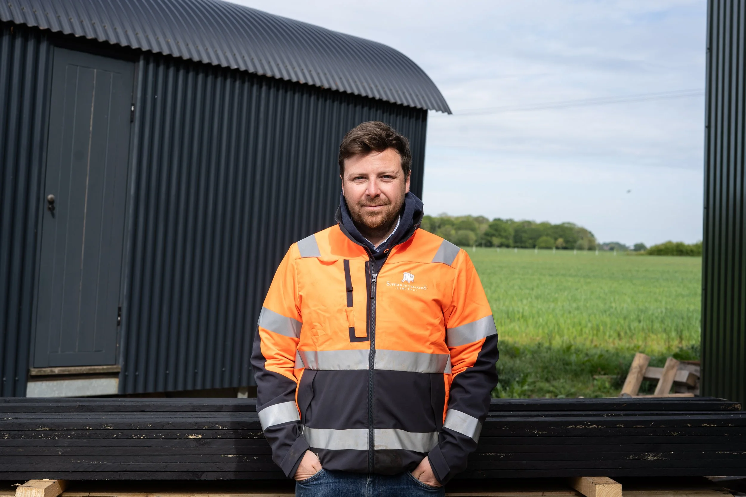 A man wearing an orange safety jacket with reflective stripes, standing outdoors in front of a black metal structure with a curved roof, and lush green fields under a cloudy sky.