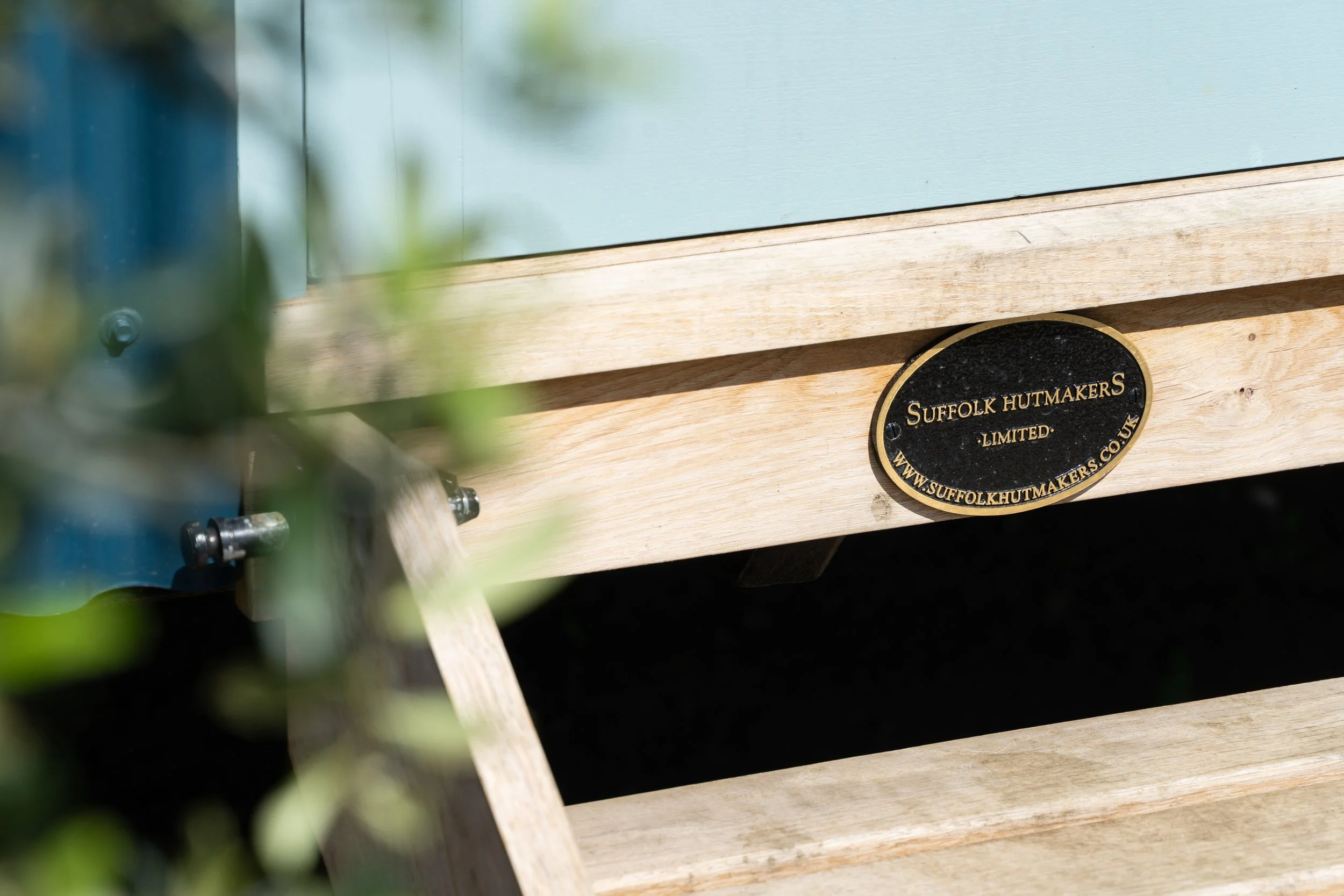 Close-up of a wooden beehive with a black and gold plaque that reads "Suffolk Hutmakers Limited" and a website URL, partially visible.