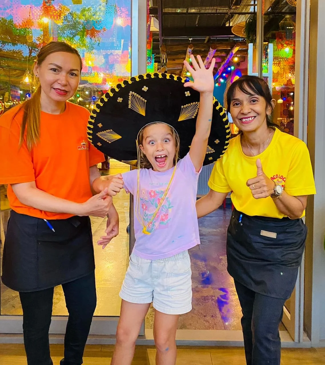 A girl wearing a colorful T-shirt, white shorts, and a large black sombrero hat, is celebrating with two women, one in an orange shirt and the other in yellow, inside a lively restaurant decorated with colorful lights.