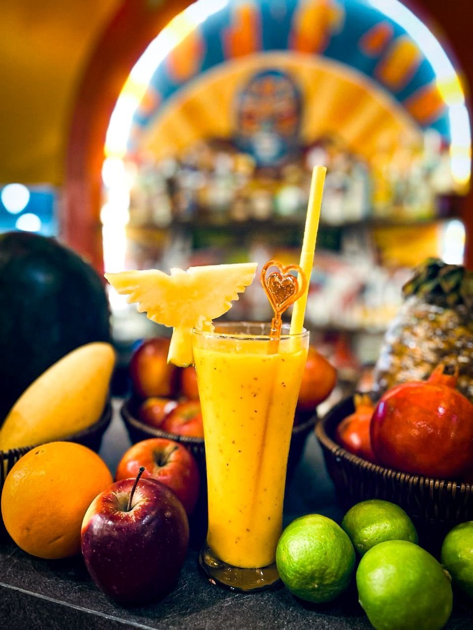 A tall glass of yellow mango smoothie garnished with a pineapple wedge and heart-shaped stir stick, surrounded by apples, pears, and limes with a colorful, blurred background.
