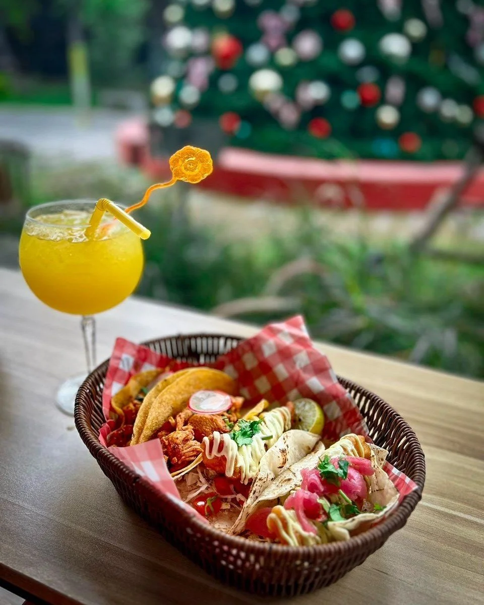 A basket of tacos and a yellow cocktail with a decorative straw and flower-shaped garnish, on a wooden table with a blurred Christmas tree in the background.