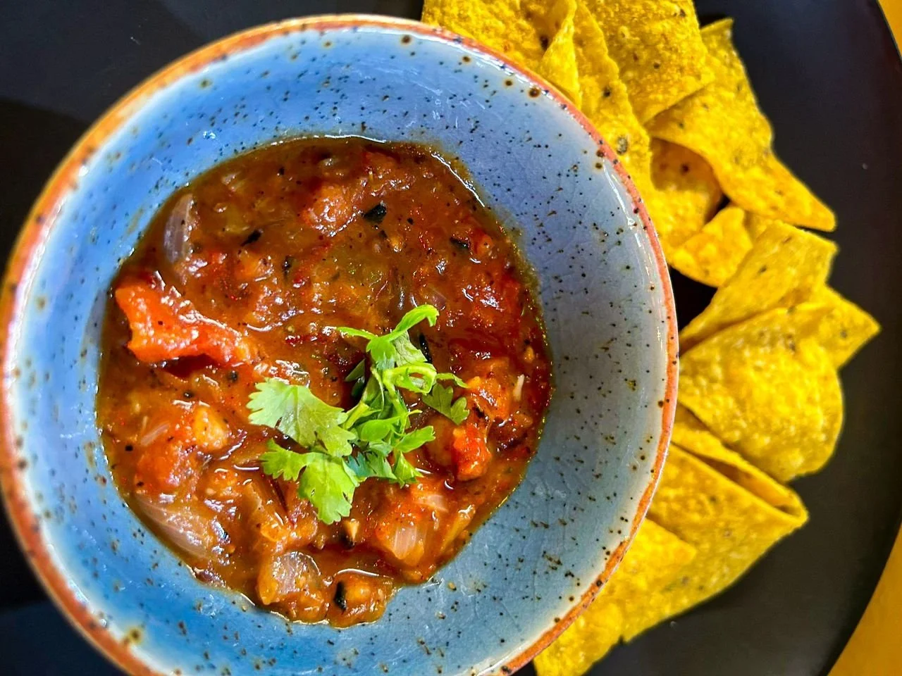 A bowl of chunky tomato-based salsa garnished with cilantro, accompanied by yellow tortilla chips on a dark plate.