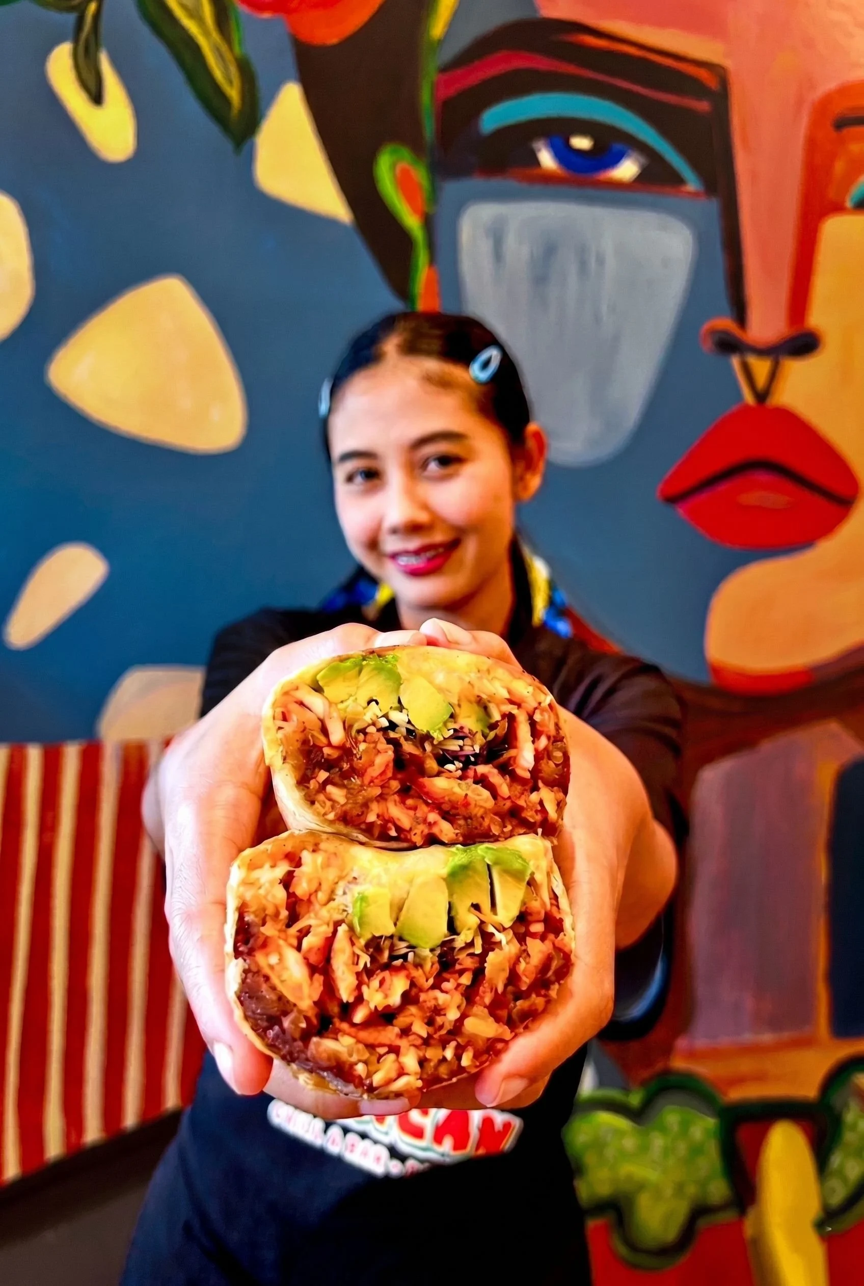 A woman with dark hair in a ponytail holds two burritos towards the camera. The burritos contain rice, vegetables, and other fillings. She is standing in front of a colorful mural with abstract shapes and a face with blue eyes, a gray mask, and red lips. She smiles and is wearing a black shirt with a logo.