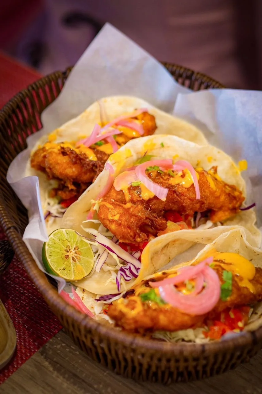 Three fish tacos with fried fish, shredded cabbage, pickled onions, and sauces in tortillas, garnished with a lime wedge, served in a basket lined with paper.