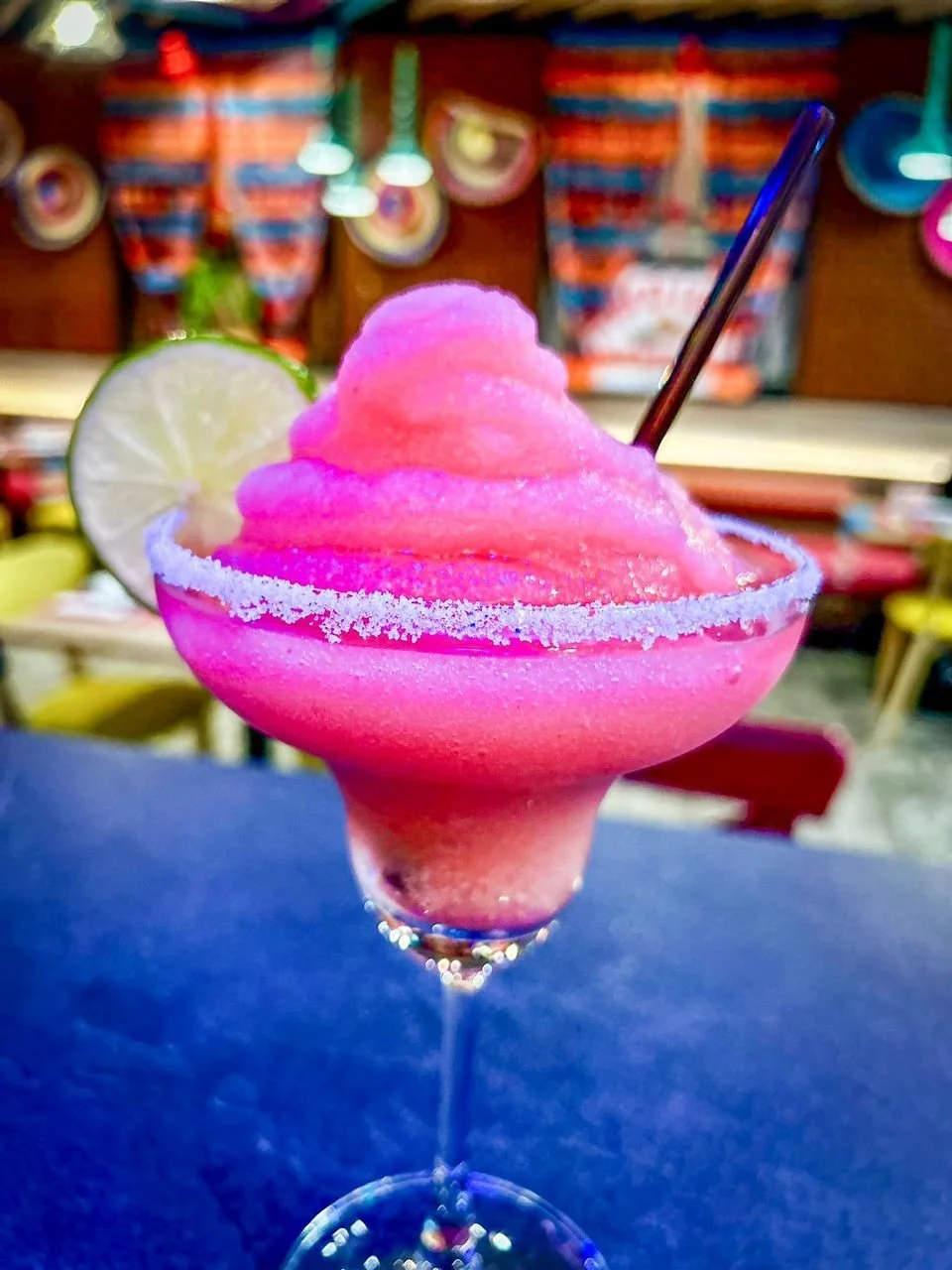 Pink frozen cocktail topped with a lime wedge and served in a margarita glass with a salted rim.