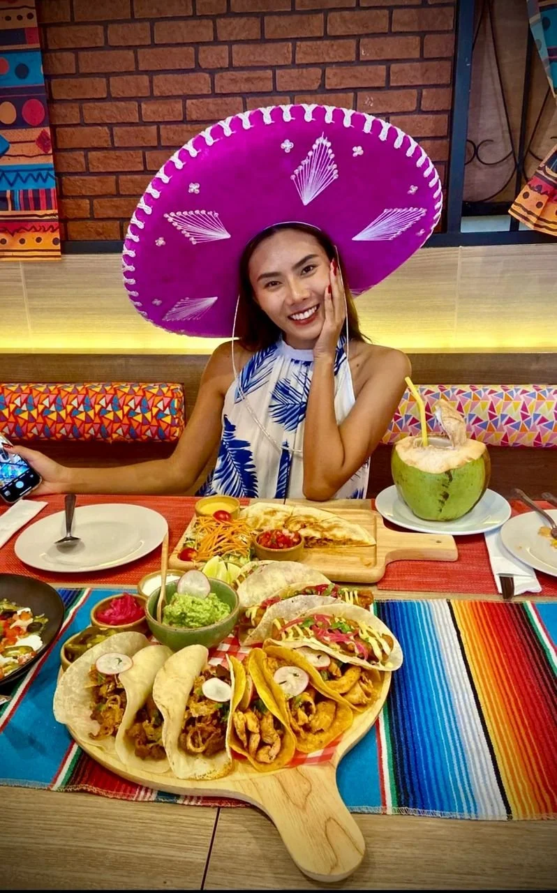 Young woman in a blue and white dress wearing a large pink sombrero, sitting at a table filled with Mexican food, including tacos, nachos, salsa, and a coconut drink, in a colorful restaurant.