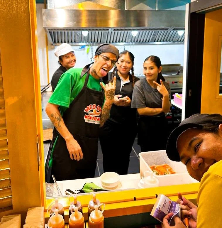 A group of four women and one man in a restaurant kitchen, smiling and making playful gestures towards the camera. There are condiments and food containers on the counter in front of them.passion for mexican cuisine