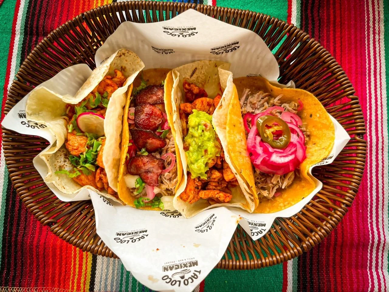 Four street tacos on a basket-lined tray with colorful woven tablecloth underneath.